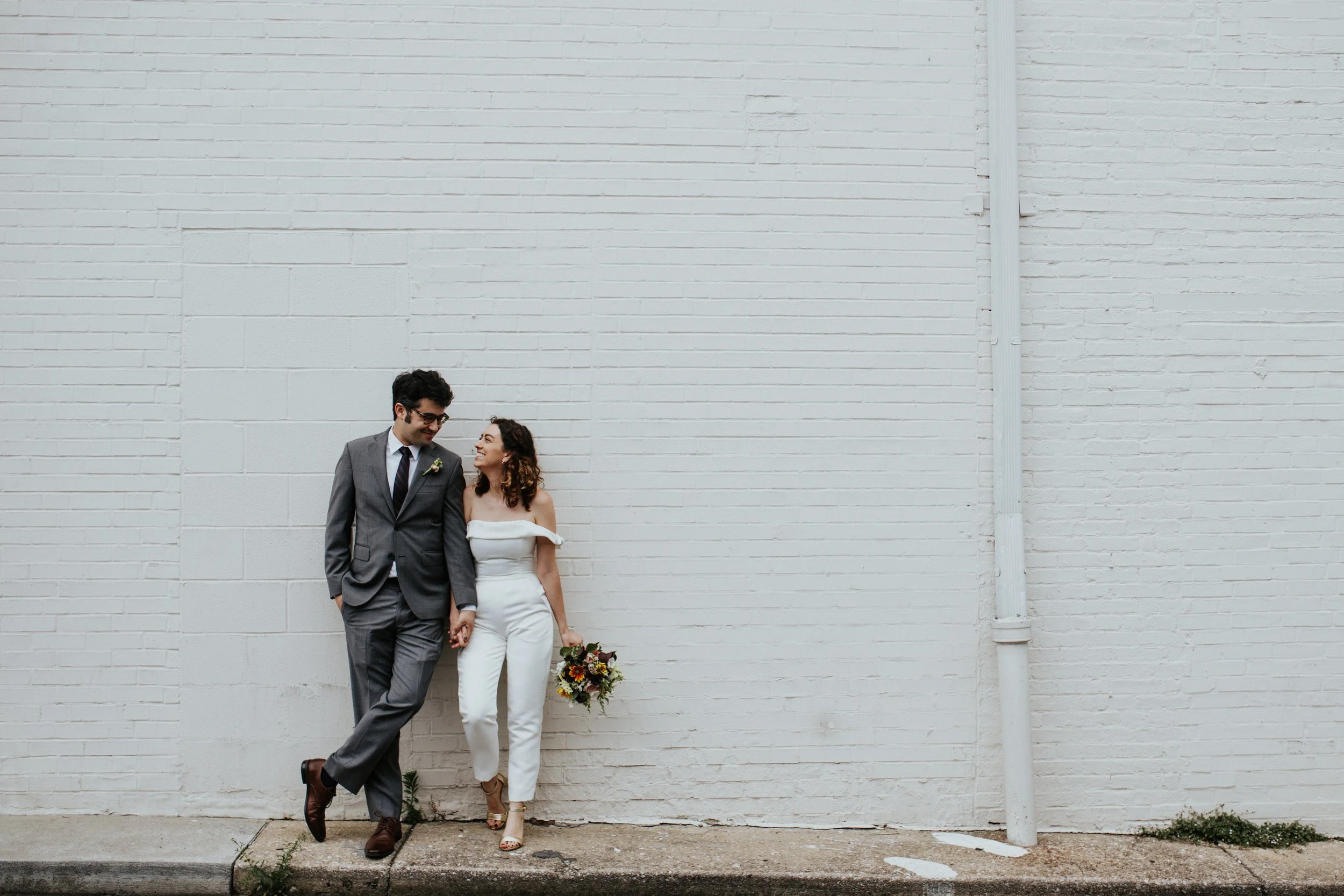 A couple dressed in wedding attire walking hand in hand along a sidewalk next to a white brick wall. The man is wearing glasses, a gray suit, and brown shoes, while the woman is in a white off-shoulder jumpsuit and holding a colorful bouquet of flowe