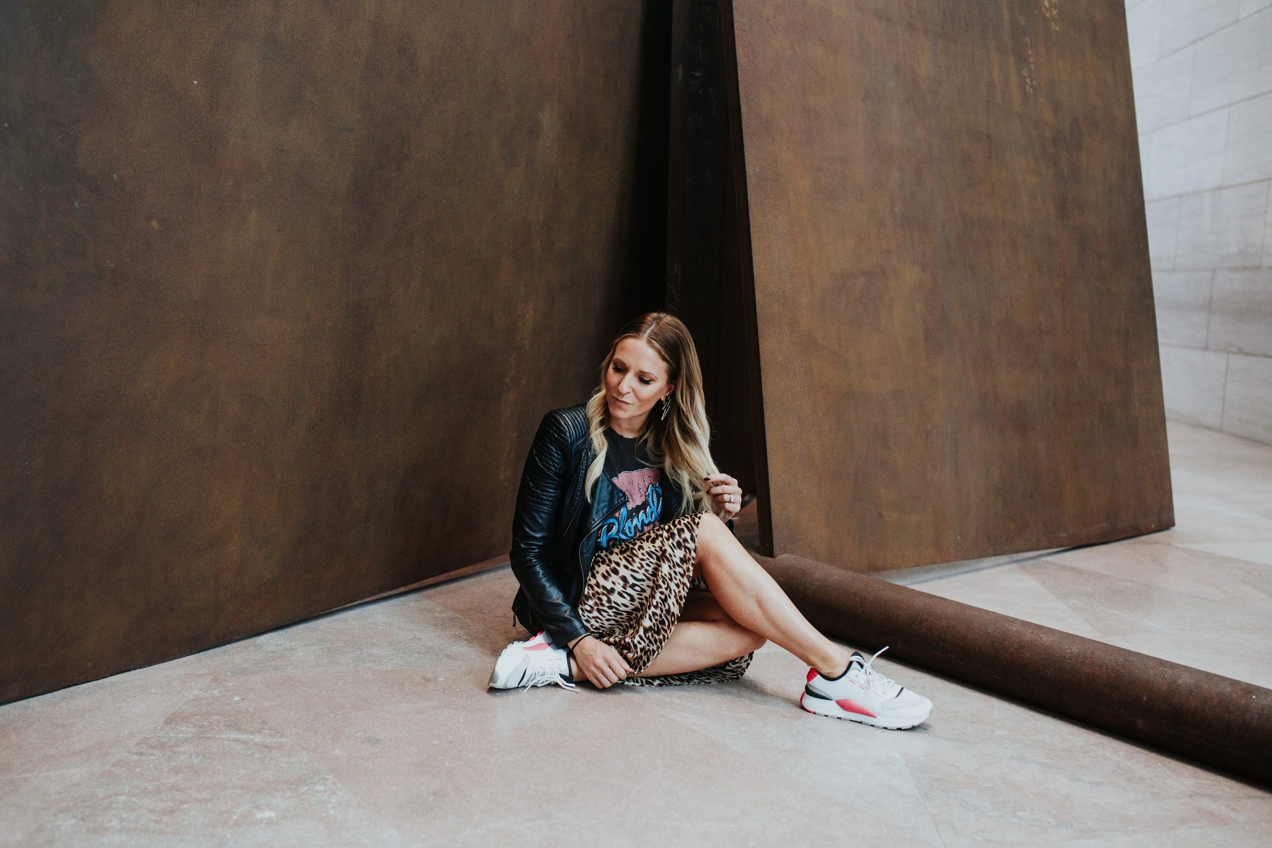 A woman sitting on the floor in front of a large brown wall, wearing a leather jacket, leopard print skirt, and white sneakers with red accents, looking down and touching her hair.