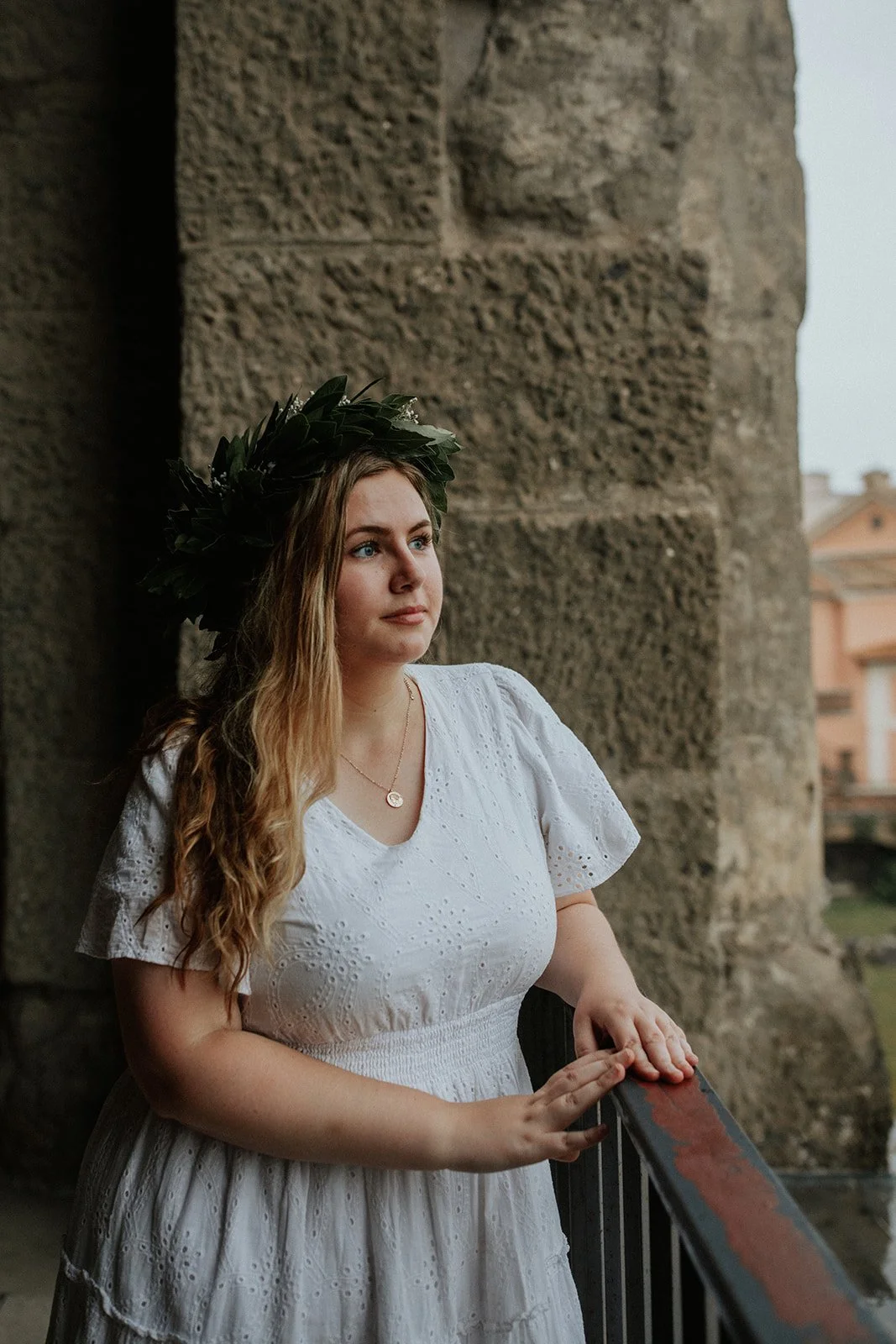 A young woman wearing a white dress and a laurel wreath stands on a balcony, looking into the distance with a contemplative expression, against a backdrop of ancient stone walls and a cloudy sky.