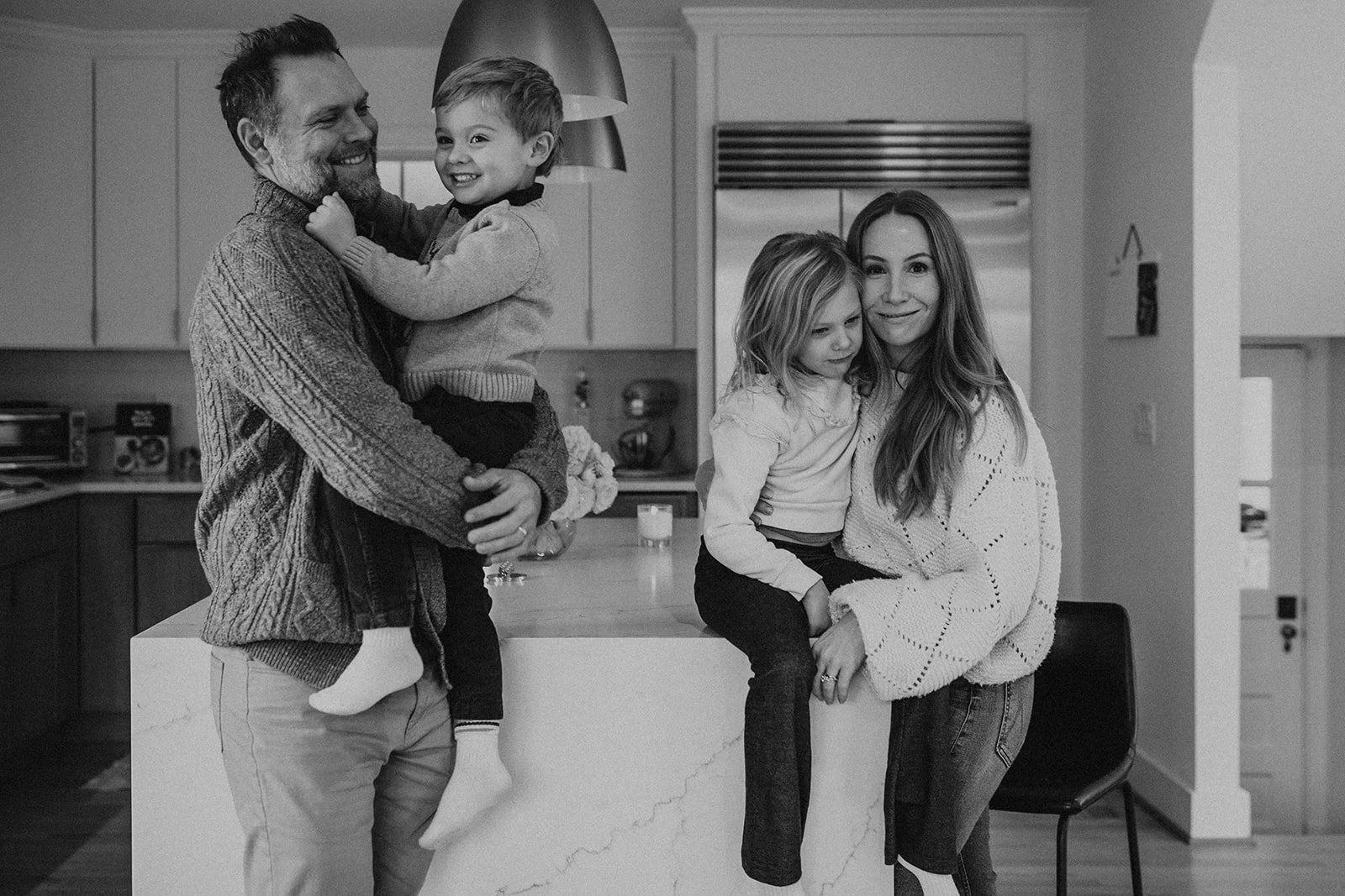 A black and white photo of a family of four in a kitchen. The father is holding a young boy who is smiling, and the mother is sitting with a young girl on a kitchen island, both smiling at the camera.