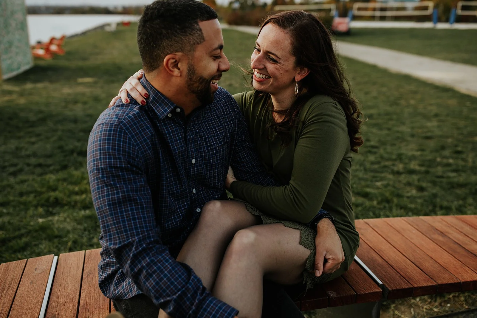 A man and woman sitting close together outdoors, smiling and looking at each other. The woman has her hand on the man's shoulder, and they appear happy.