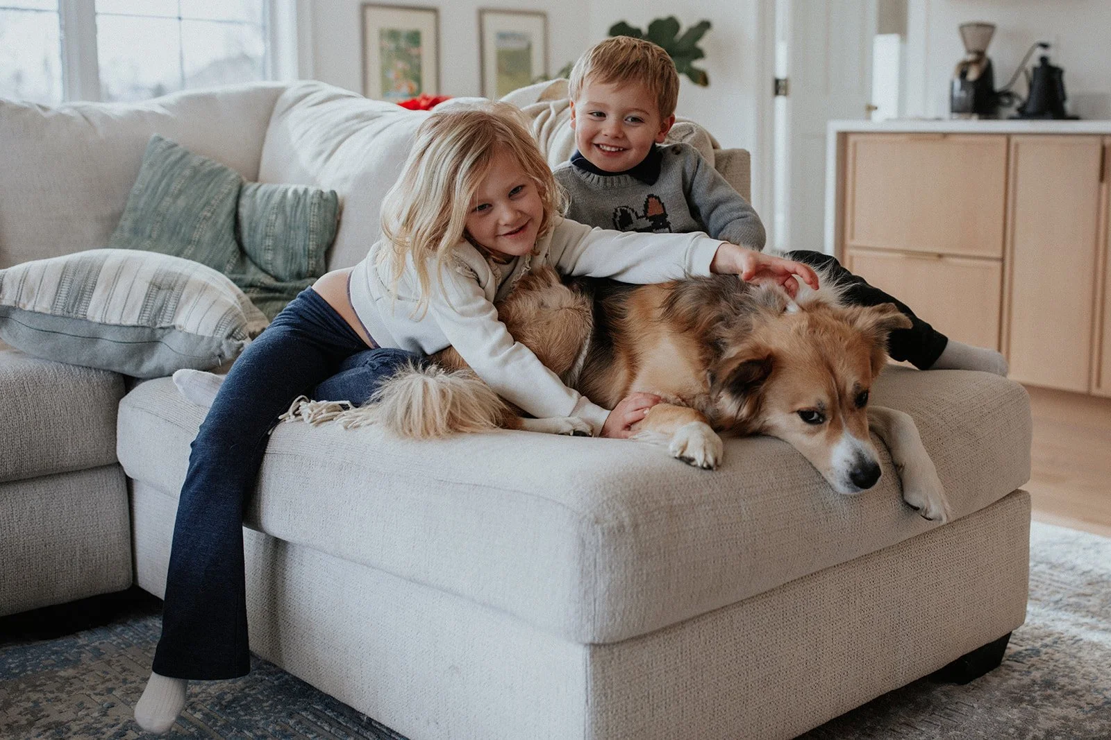 Two children and a dog lying on a beige couch in a cozy living room.