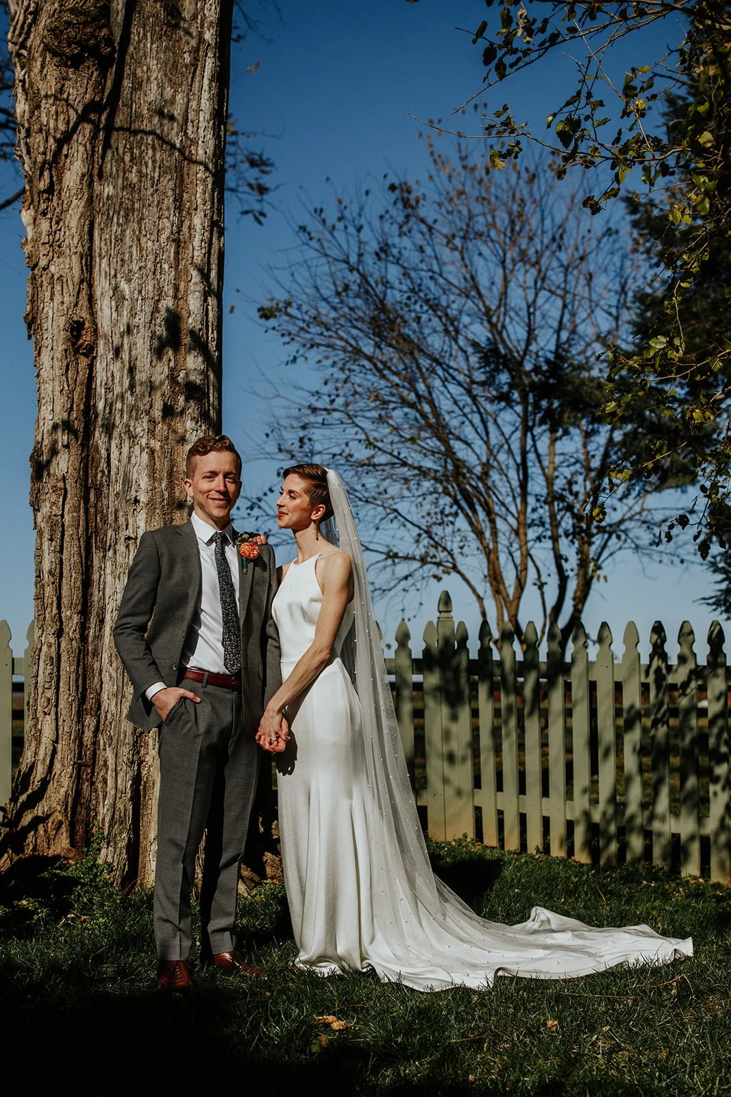 A couple dressed in wedding attire standing outdoors by a large tree and white picket fence, holding hands, with a clear blue sky in the background.