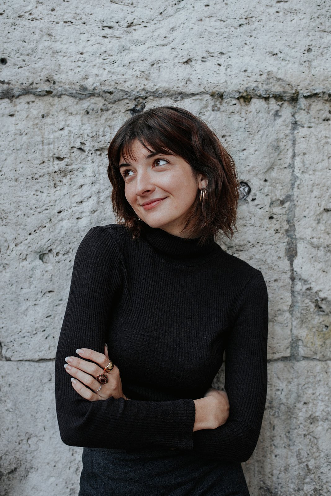 A young woman with shoulder-length dark hair, wearing a black turtleneck sweater, standing against a stone wall, smiling softly and looking to her left.