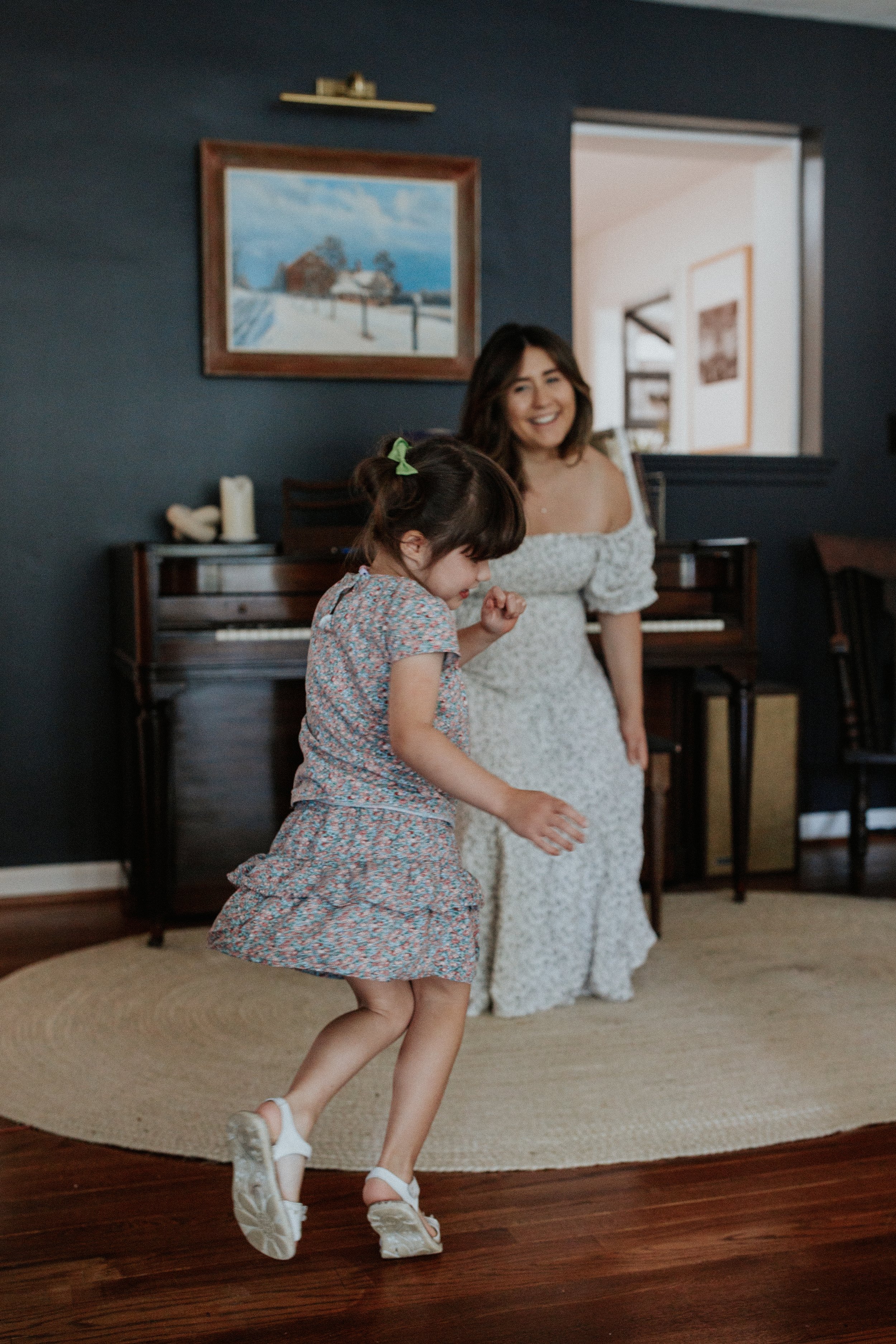 A young girl is dancing with a woman in a living room. The girl is wearing a floral dress and white shoes, and the woman, possibly her mother, is wearing a white dress with a floral pattern. They are smiling and appear to be enjoying a fun moment tog