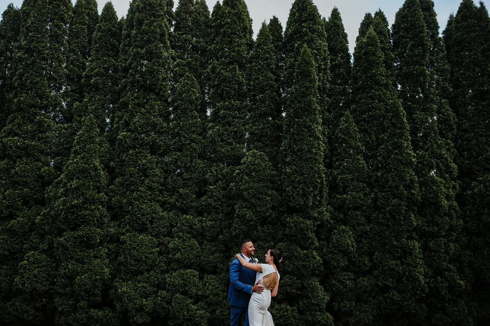 A bride and groom standing close, smiling and looking into each other's eyes, in front of a large, dense green hedge or row of trees, during a wedding celebration.