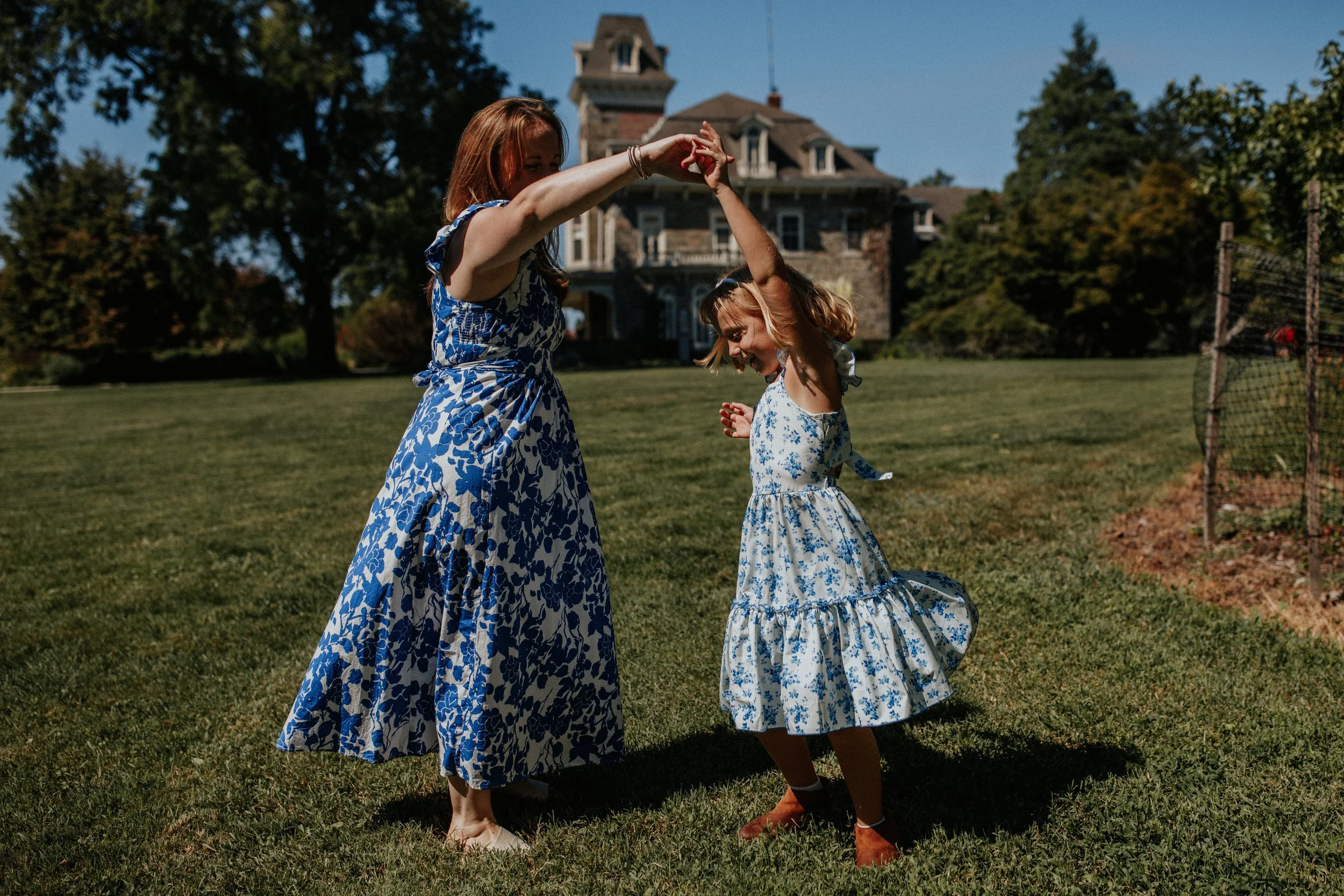 Two women dancing outside on a grassy lawn with a large stone house in the background. Both women are wearing matching blue and white floral dresses.
