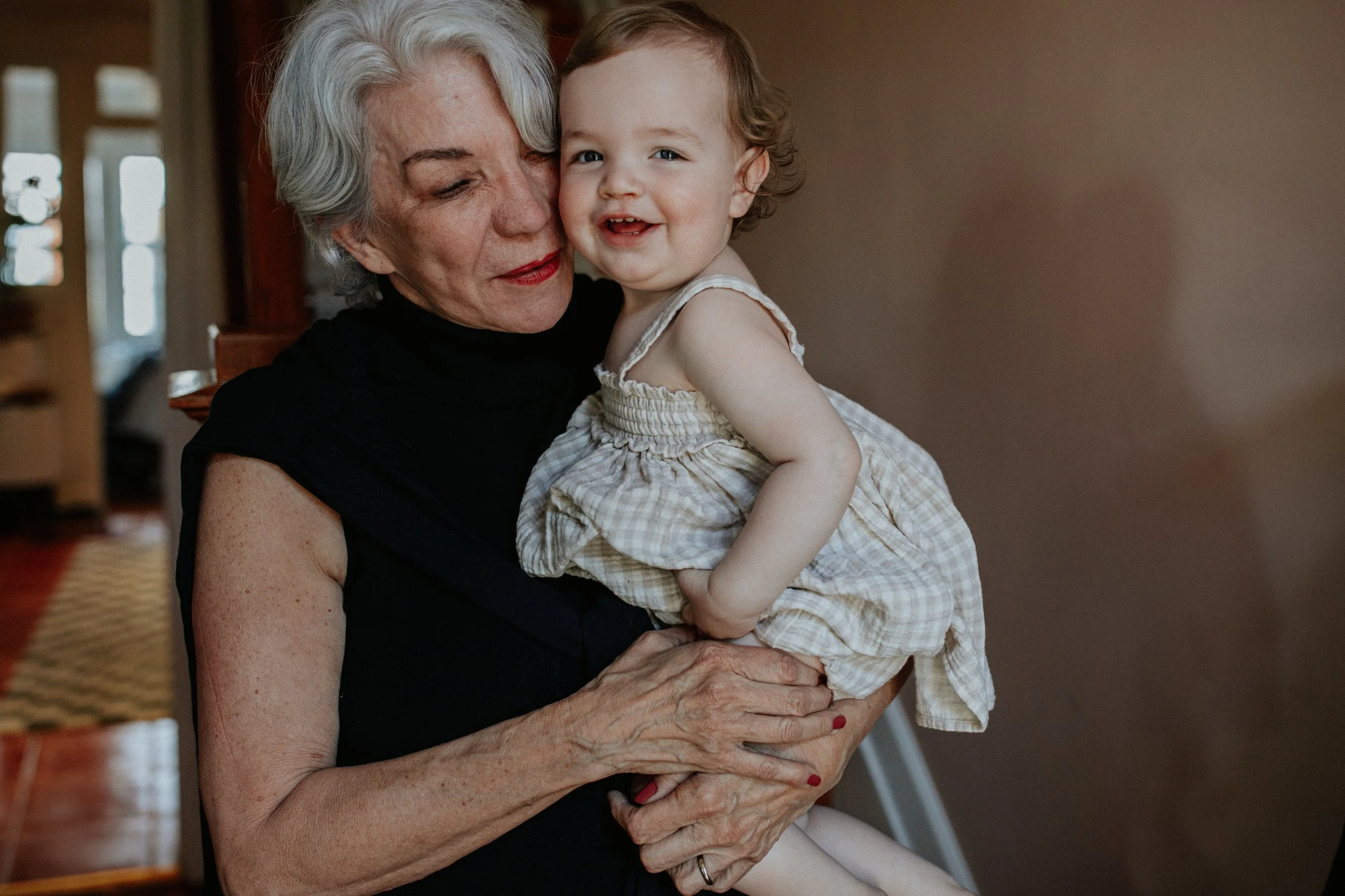An elderly woman with gray hair holding a young girl with curly hair, both smiling and close to each other inside a home.