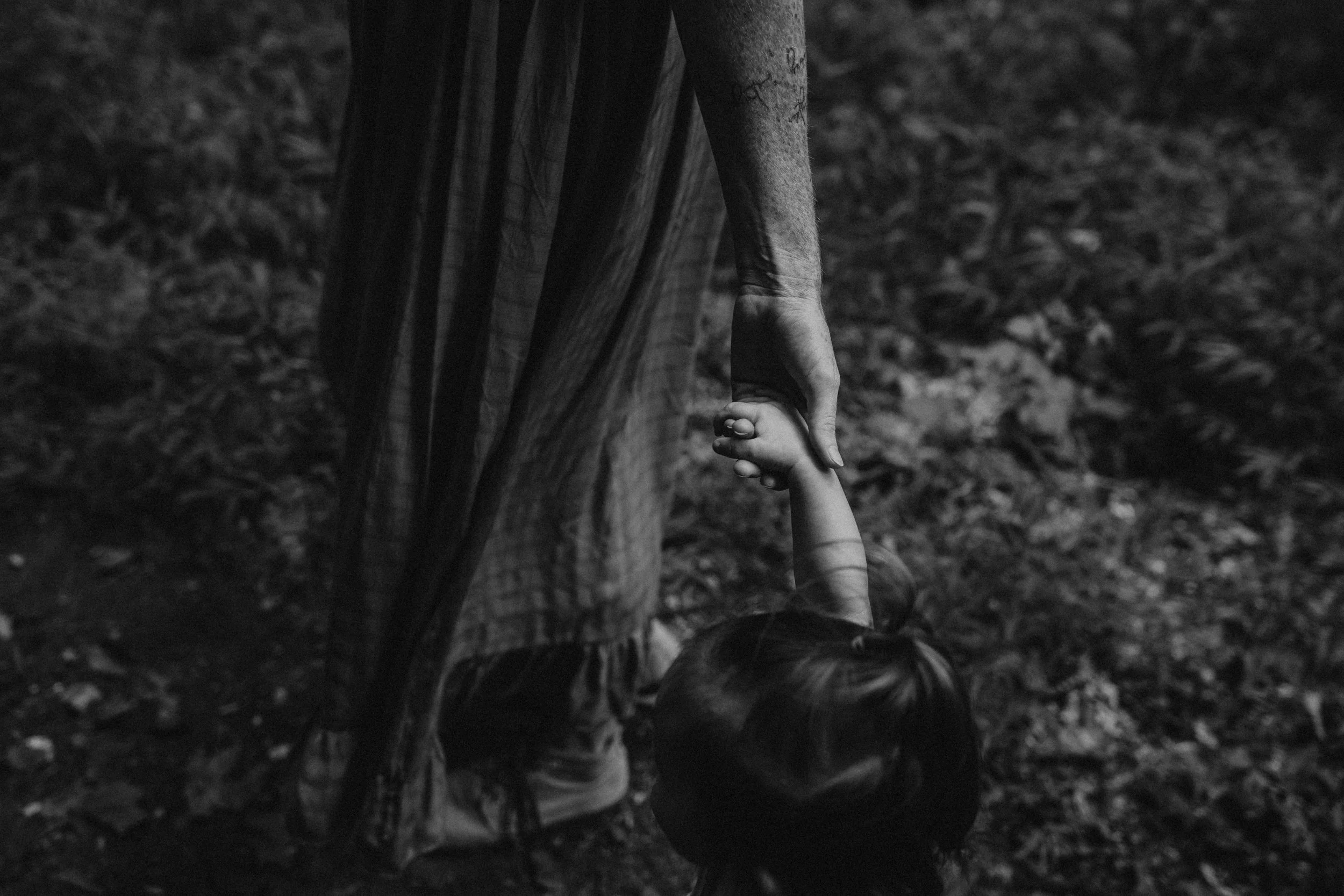 A black and white photograph showing an adult and a child holding hands while walking outdoors. The image focuses on their hands and legs, with a background of blurred natural ground and trees.