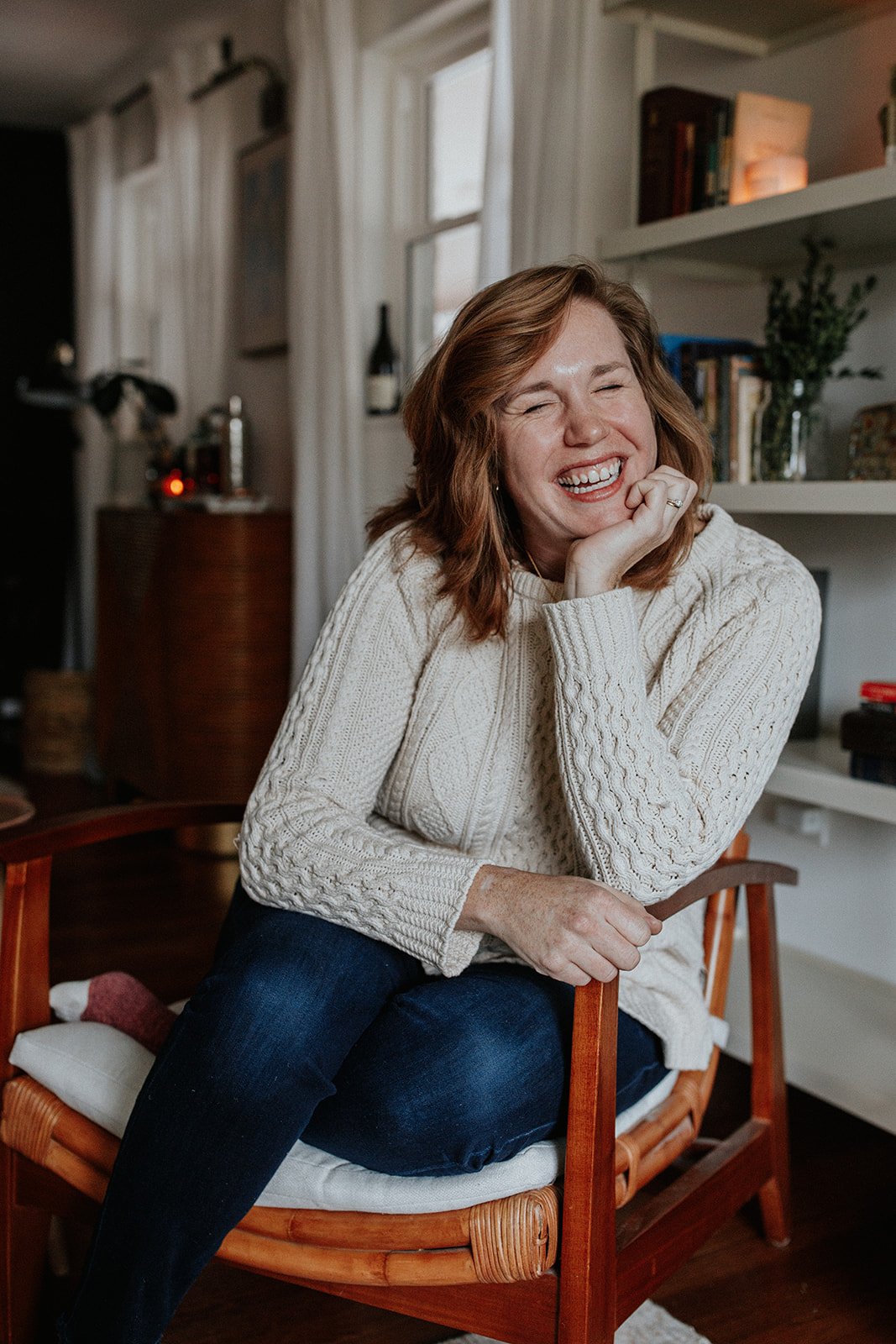 A woman with red hair wearing a cream cable-knit sweater and dark jeans, sitting on a wooden chair with a white cushion, laughing with her hand on her chin in a cozy living room with bookshelves and window behind her.