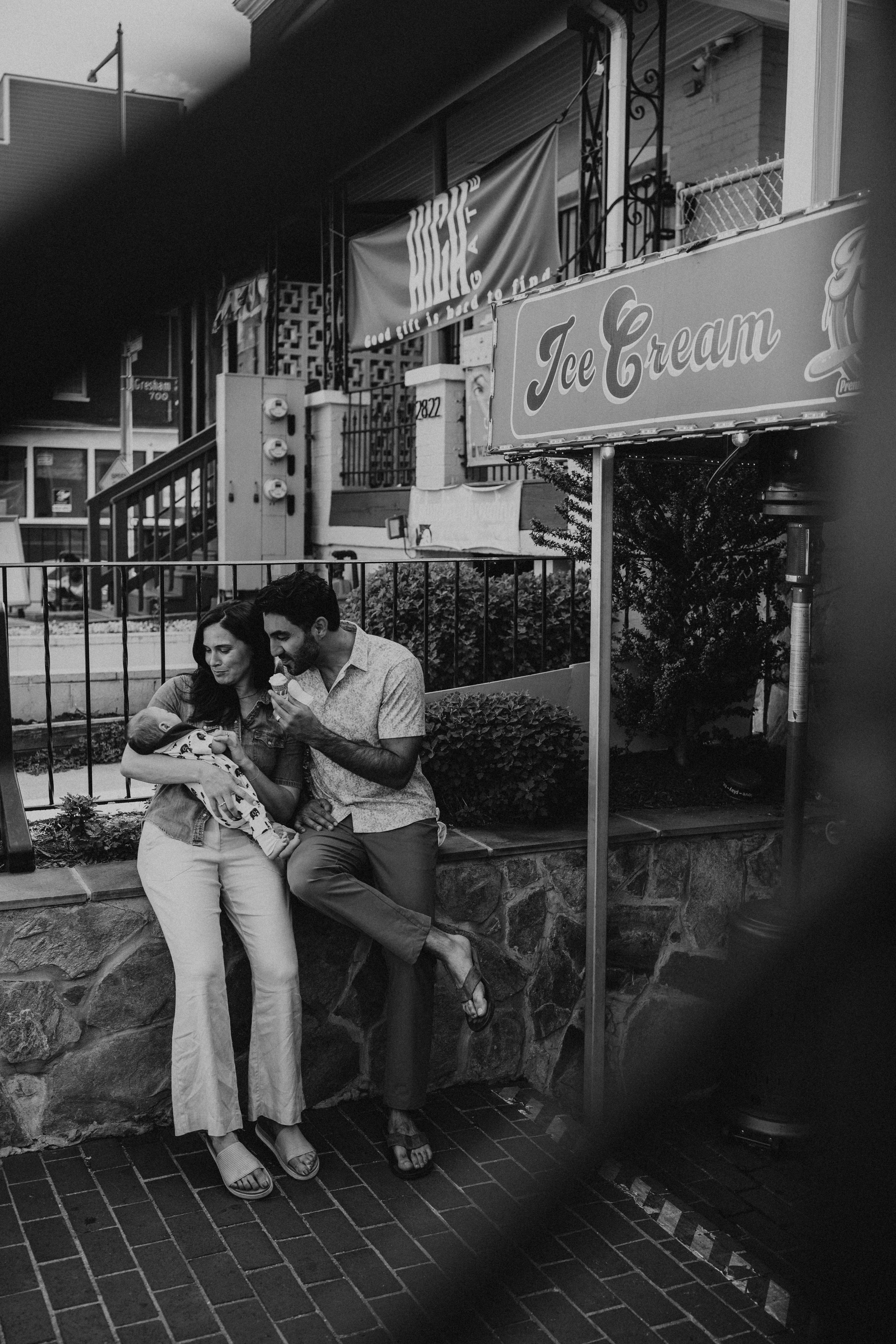 A man and woman sitting on a low stone wall, holding a baby wrapped in a blanket, in front of a fenced outdoor cafe or restaurant with a sign that says "Ice Cream." The scene is in black and white and appears to be in an urban area.