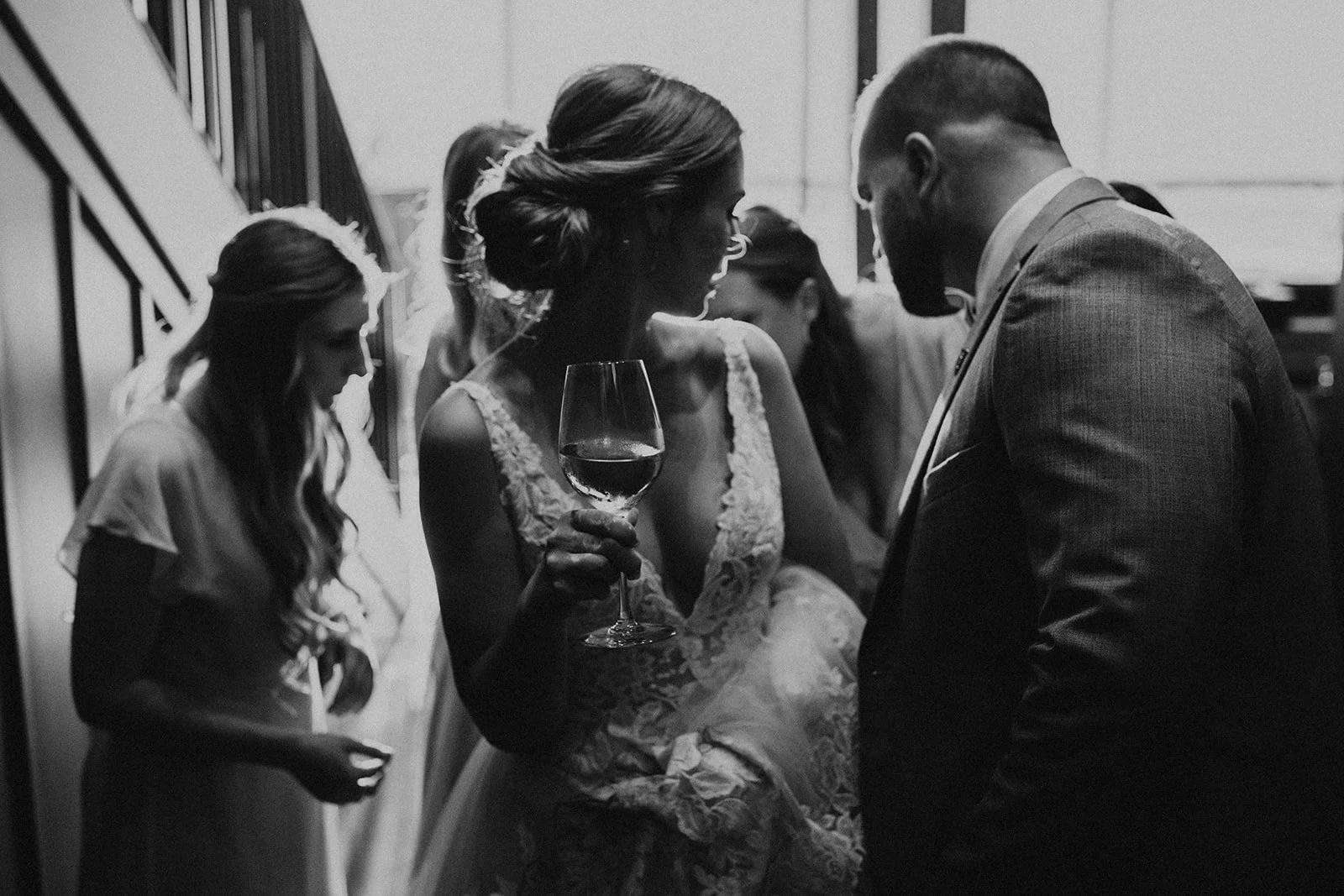 Black and white photo of a woman at a wedding reception holding a glass of red wine, surrounded by other women and a man in a suit.