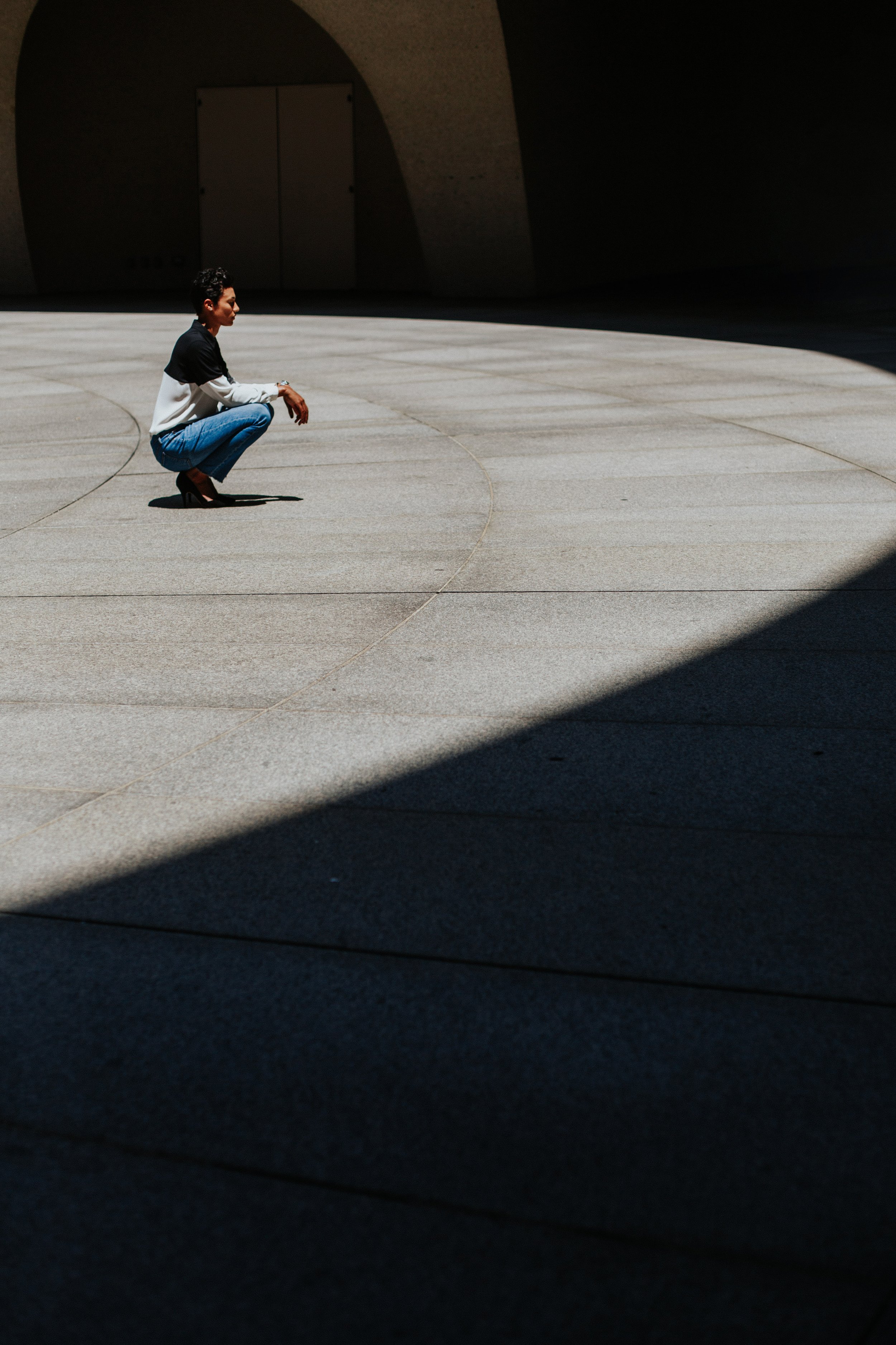 A woman squatting on a large, open, concrete plaza with a dark shadowed area on the right side and an arched structure with a closed door in the background.