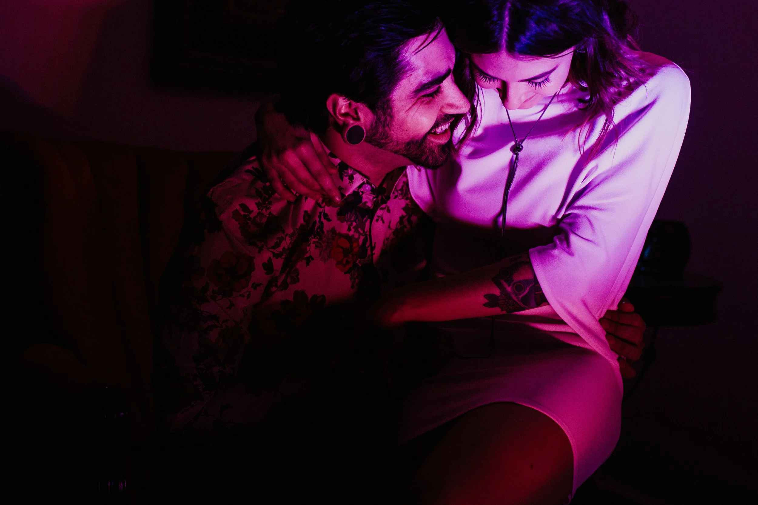 A man and woman sharing a close, intimate moment with their foreheads touching, in a dimly lit space with pink and purple lighting.