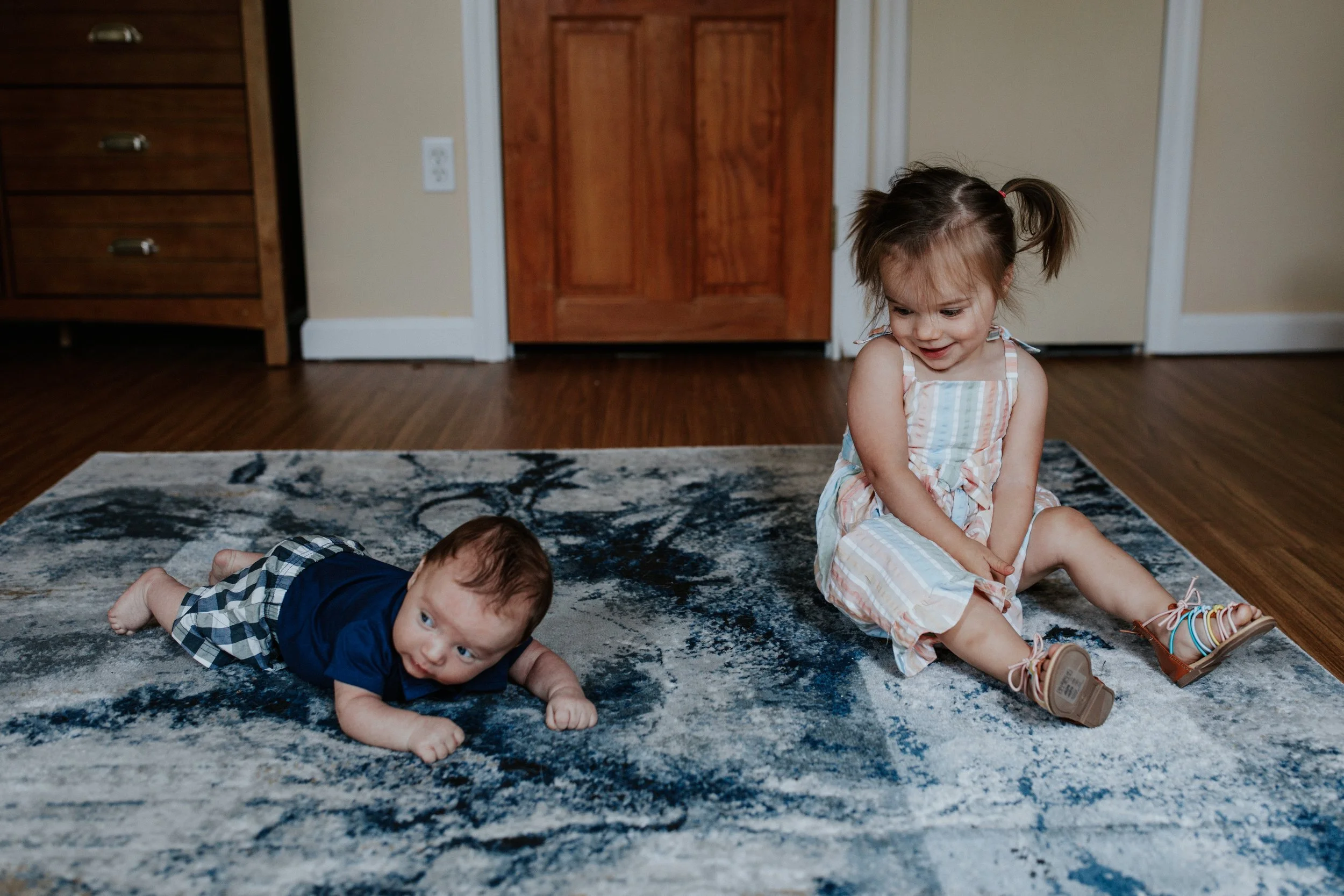 Two young children playing on a rug in a room, one girl sitting upright and smiling, and one boy lying face down on the rug.