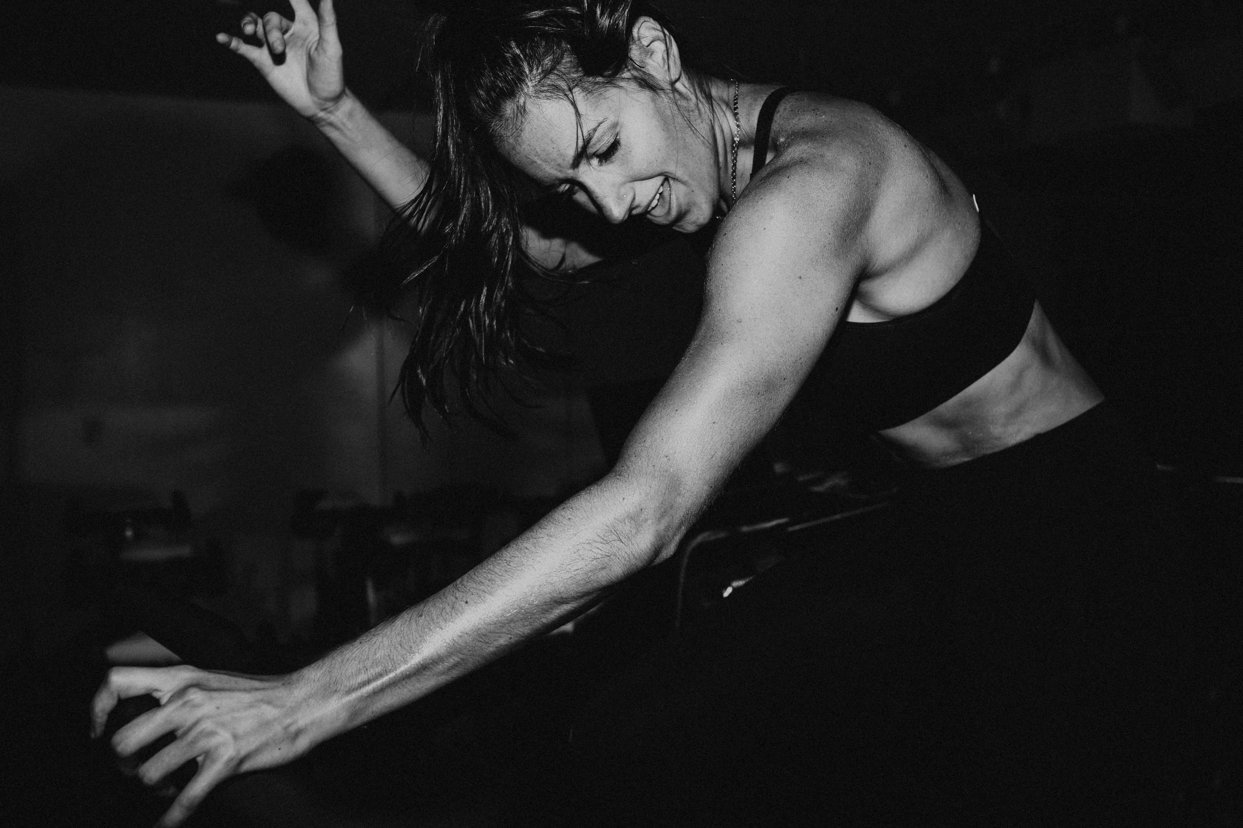 A woman with dark, wet hair in a black tank top appears to be in the middle of a dance or exercise, with her eyes closed and a joyful expression, against a dark background.