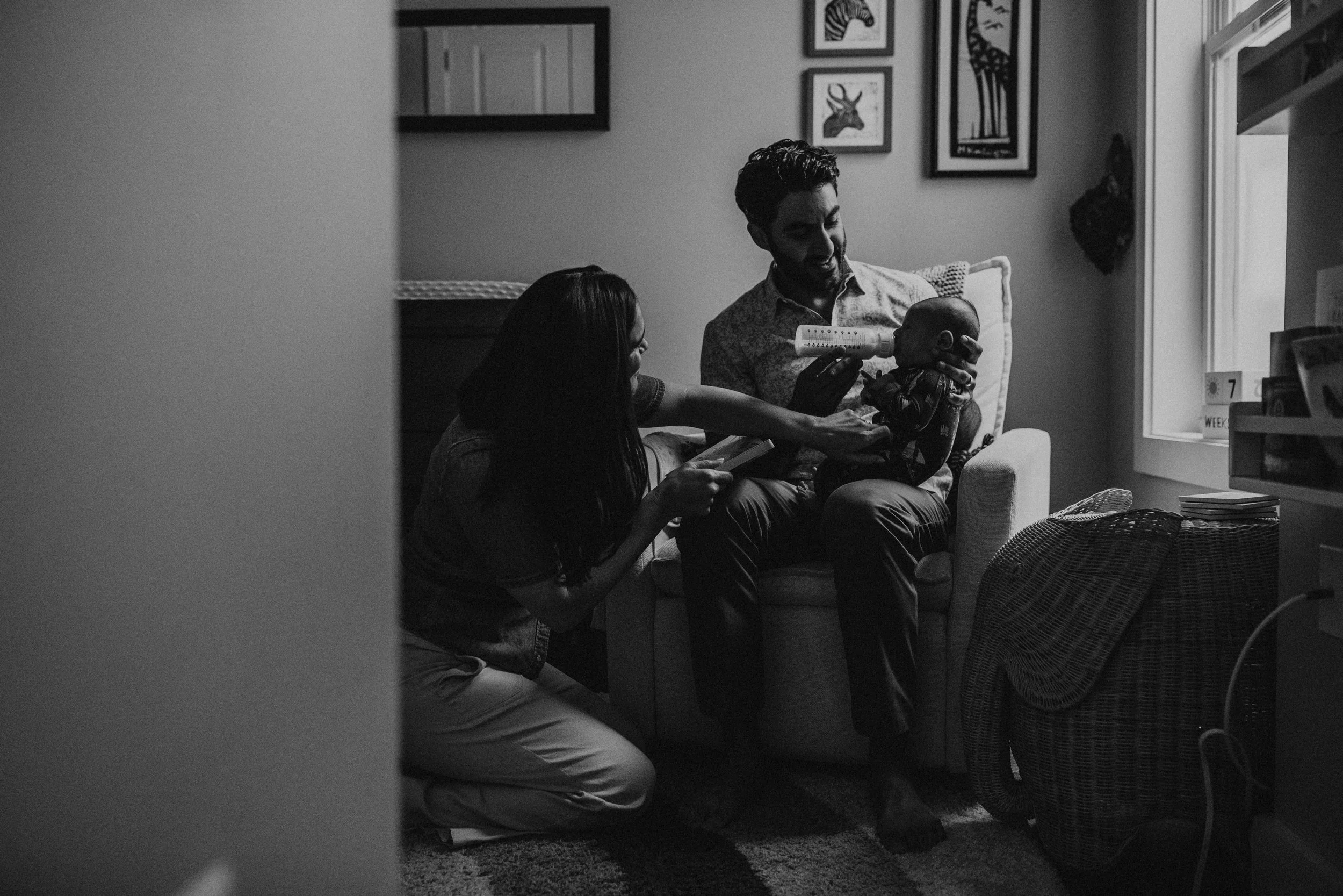A man and woman sitting on a couch in a living room, feeding a baby. The woman is holding a smartphone, and the man is feeding the baby with a bottle. The room has framed animal pictures on the wall, a window, a fan, and a calendar.