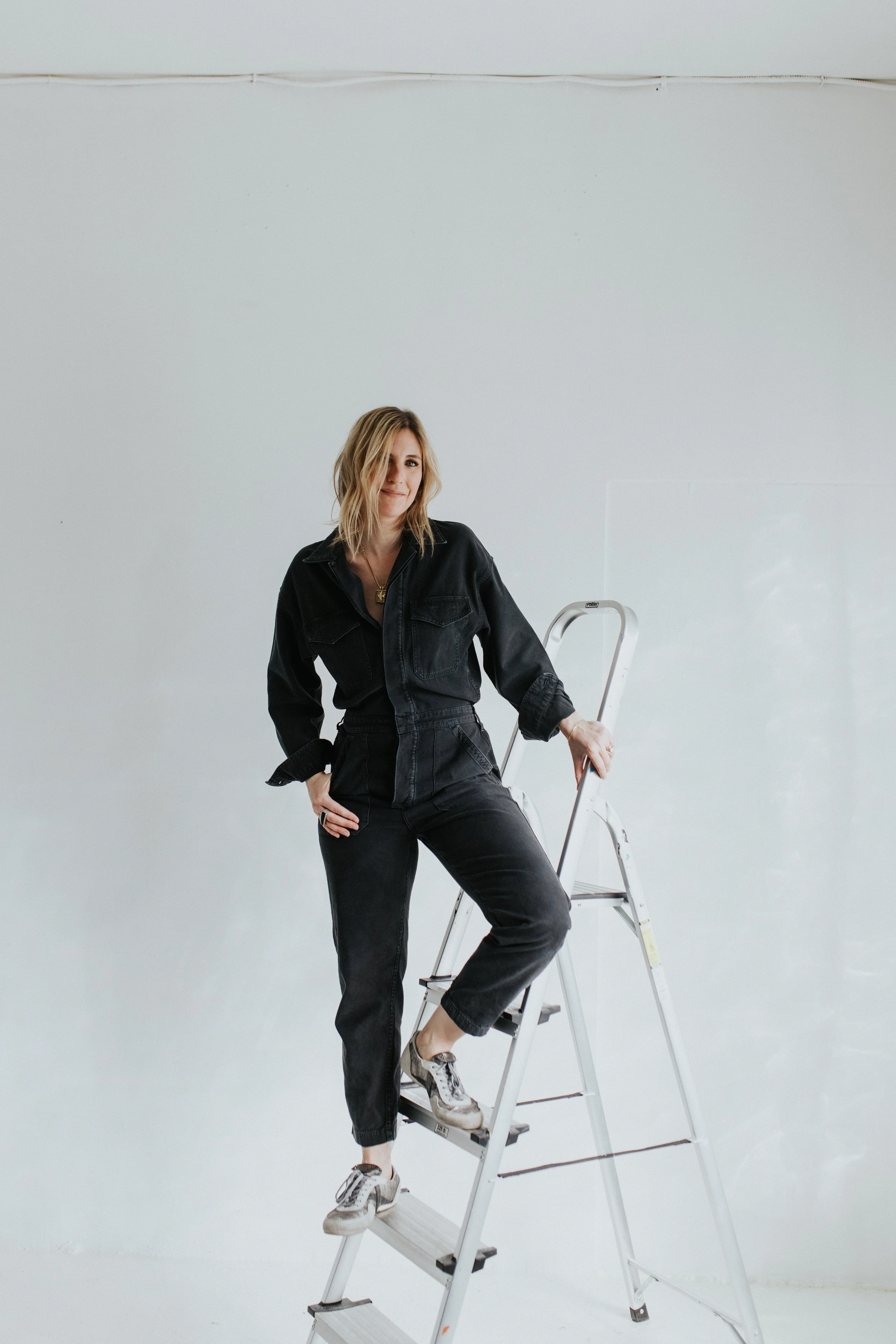A woman with blonde, wavy hair poses on a white ladder against a plain light gray wall. She is wearing a black denim jumpsuit, sneakers, and a necklace, with one hand resting on her hip and the other on the ladder.
