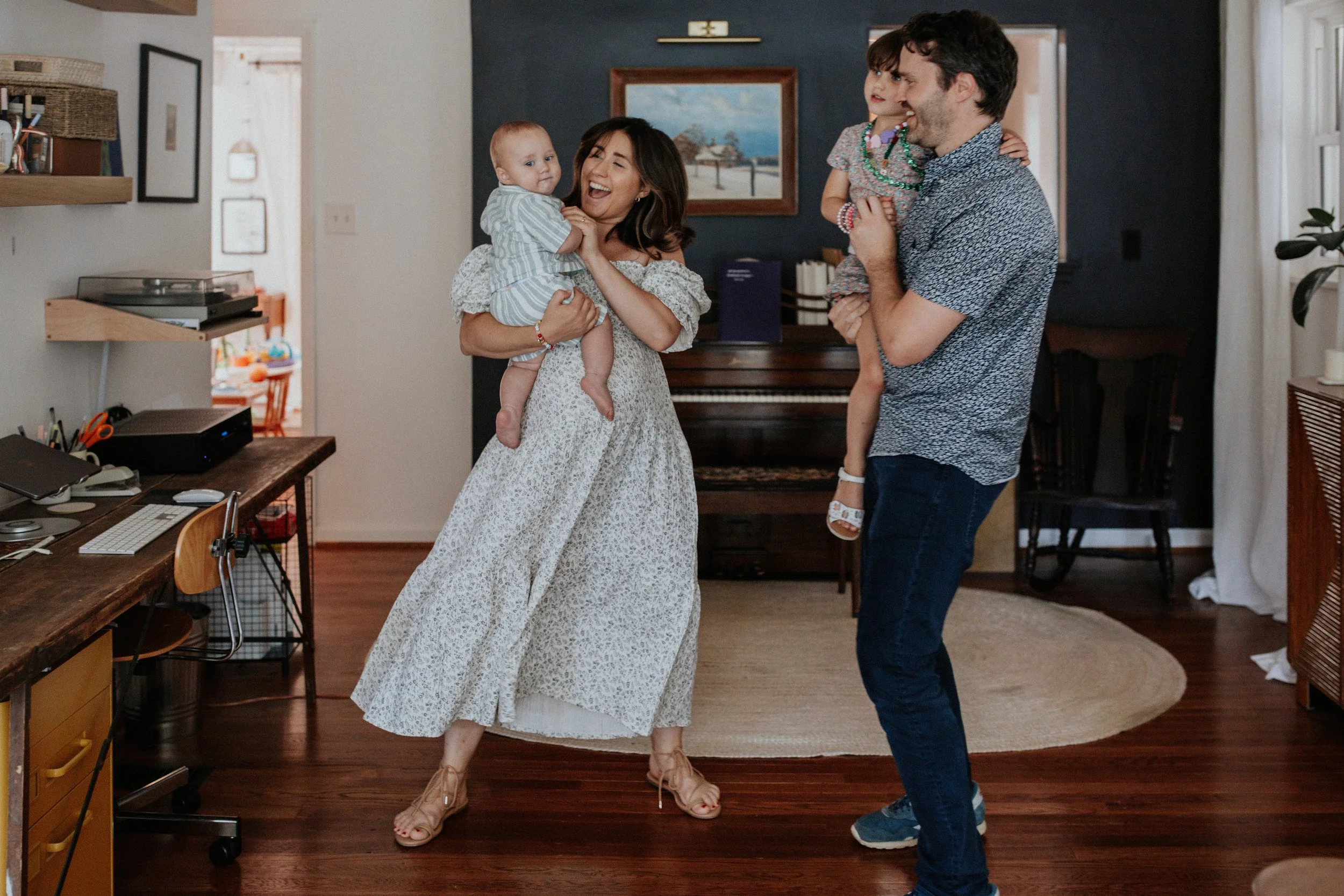 A family of four, including a woman, a man, and two young children, enjoying a happy moment indoors. The woman is holding a baby girl and smiling, while the man is holding an older child and smiling. The setting appears to be a living room with a pia
