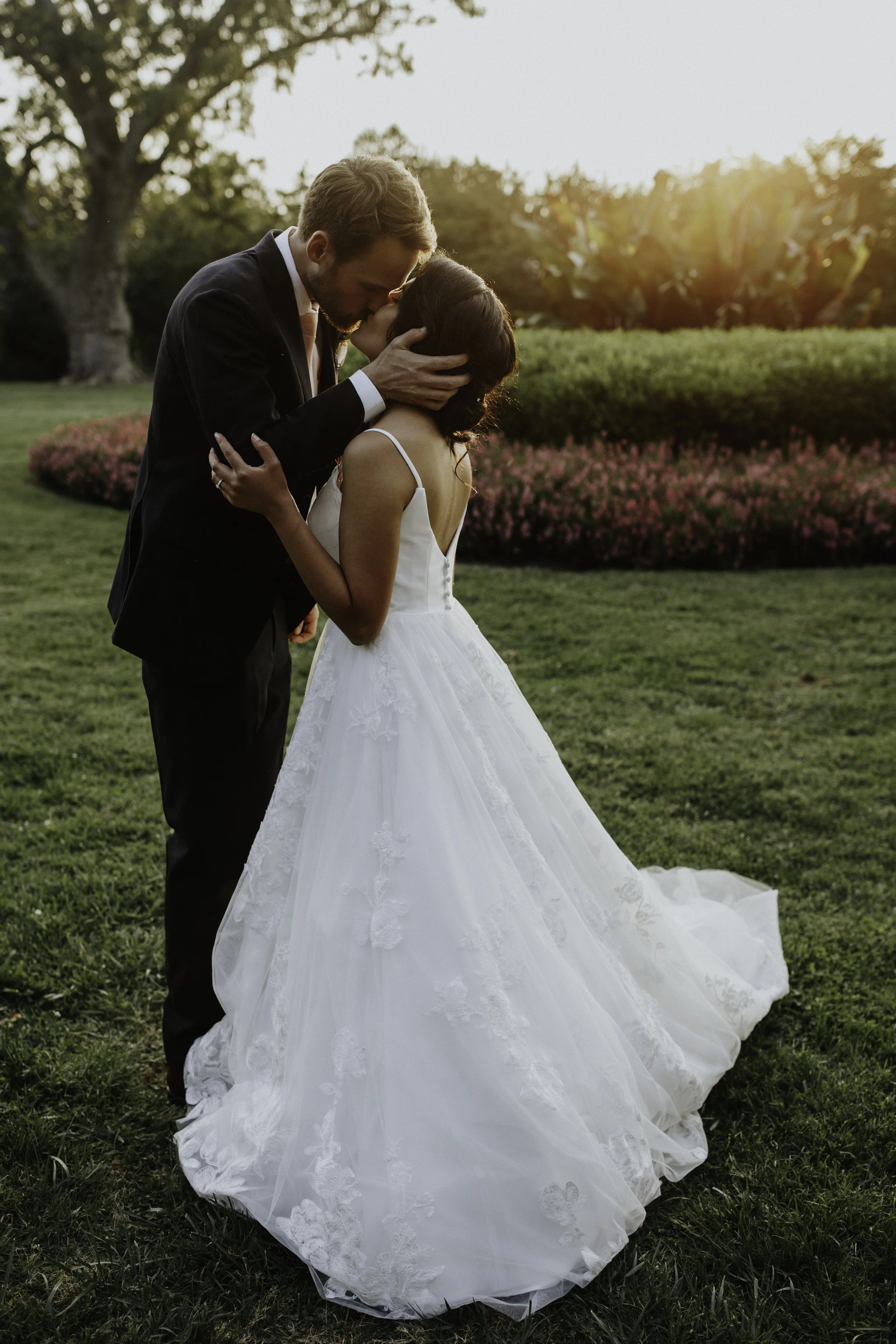 A couple, dressed in wedding attire, sharing a kiss outdoors during sunset with trees and bushes in the background.