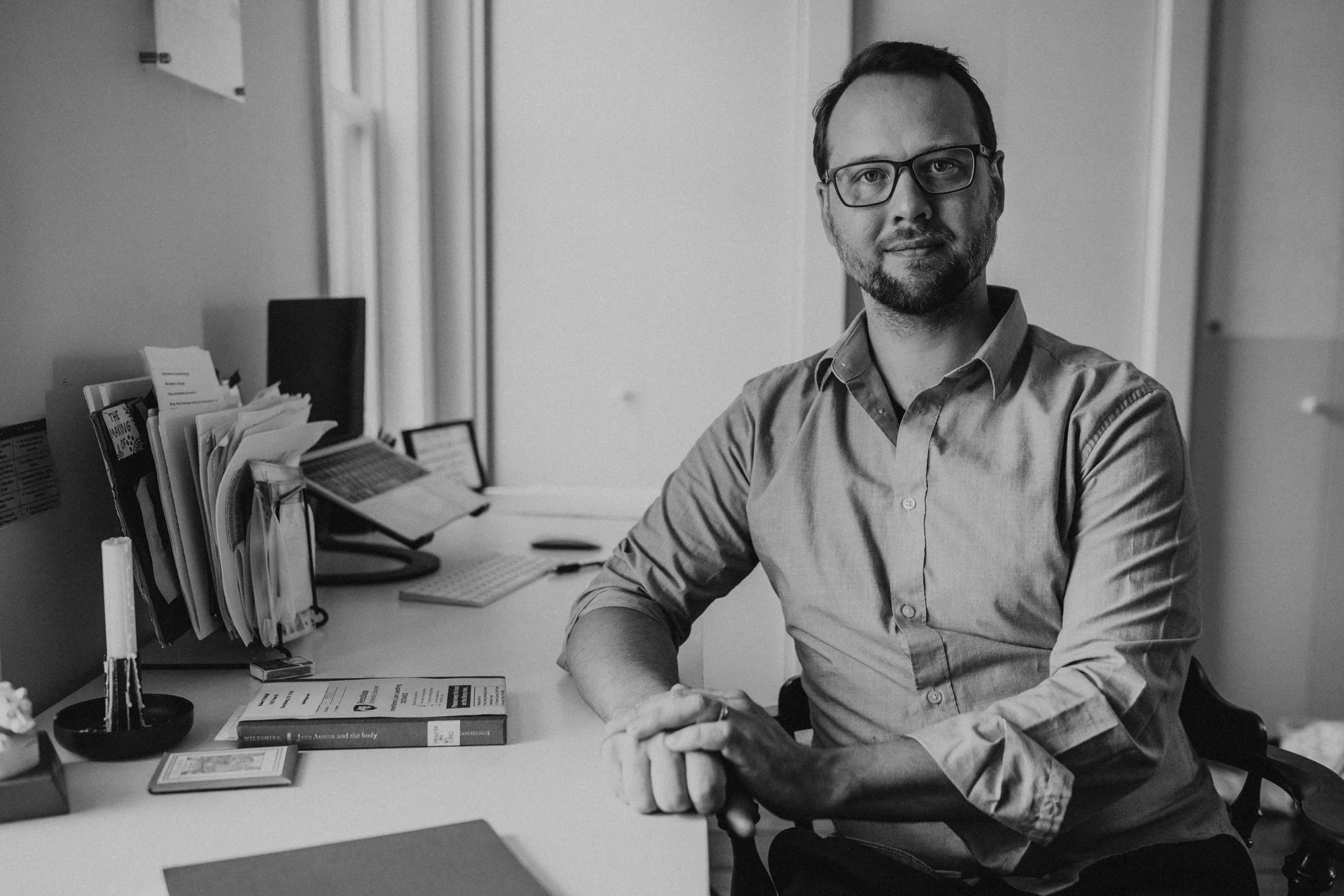 A black-and-white photo of a man with glasses and a short beard sitting at a desk in an office. He is wearing a button-up shirt and looking slightly to the side.