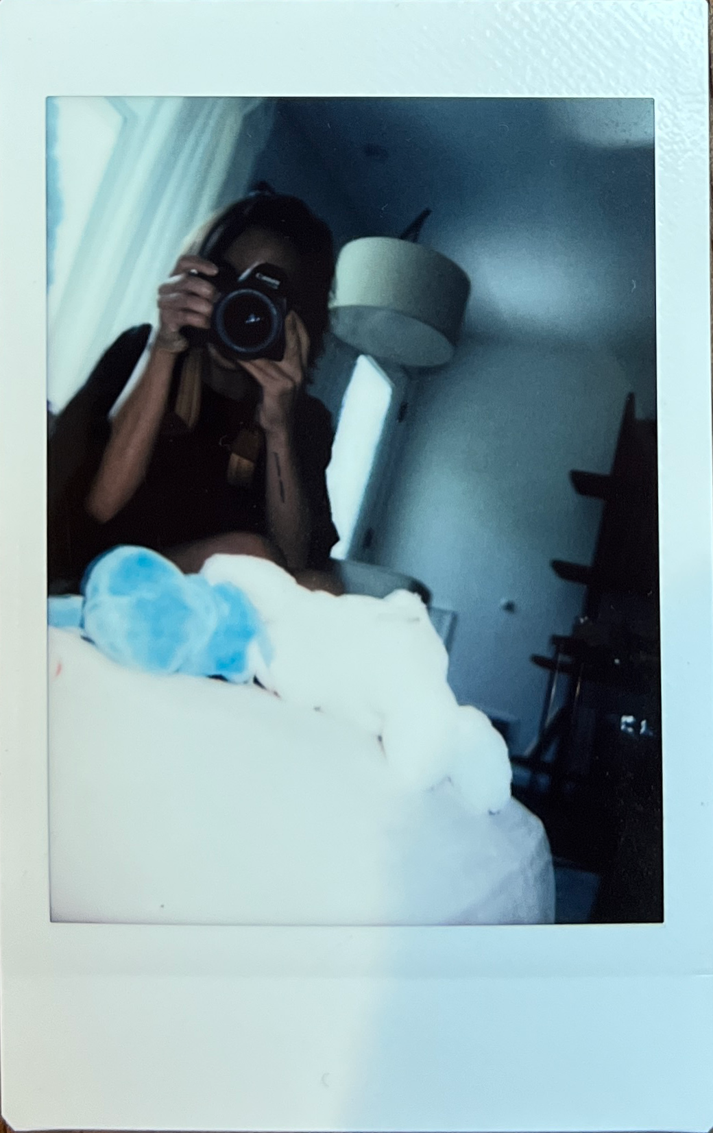 A person taking a mirror selfie in a dimly lit room, holding a camera with a large lens, with a bed and blue stuffed toy in the foreground, and a ceiling light fixture in the background.