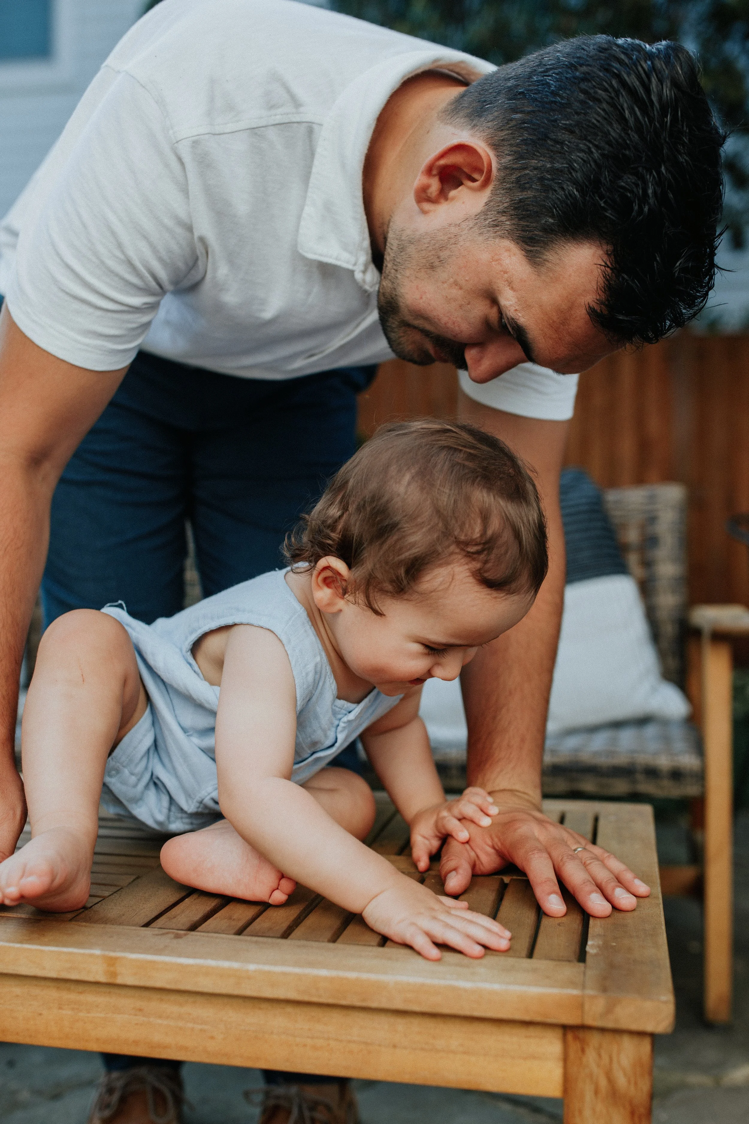 Man and small child on a wooden deck, leaning over and placing hands on the surface, engaged in an outdoor activity.