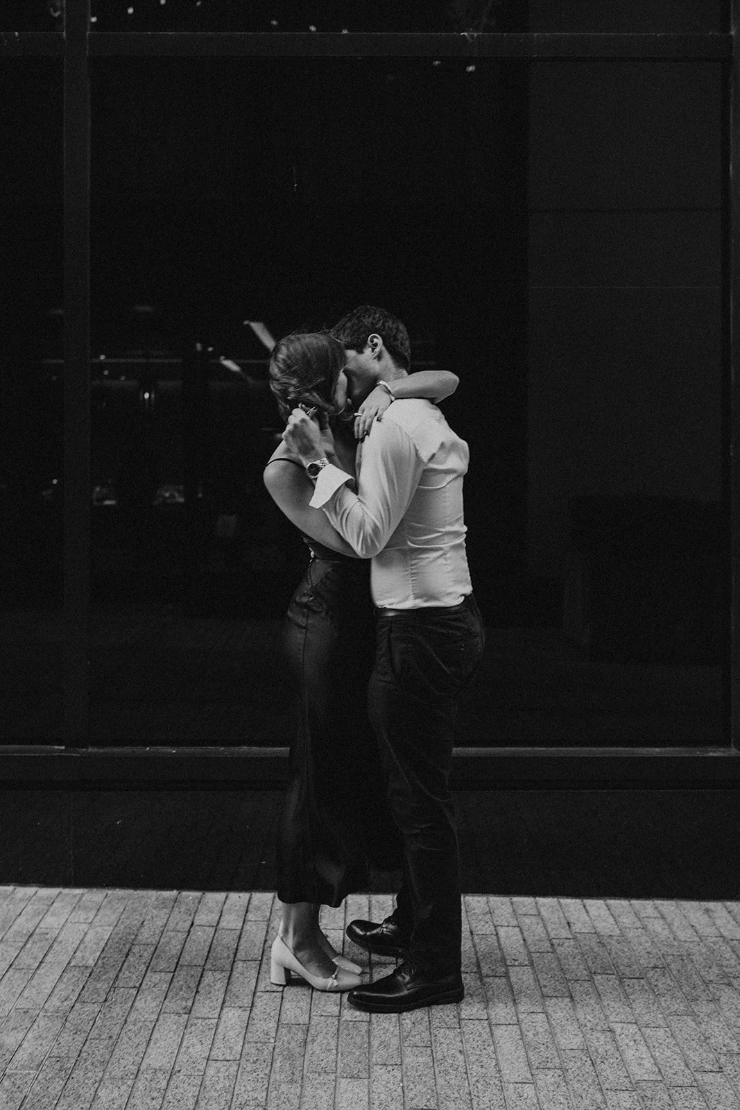A black and white photo of a couple sharing a kiss, with the woman wearing a skirt and high heels, and the man wearing a dress shirt and pants, standing outside on a brick sidewalk in front of a dark wall.