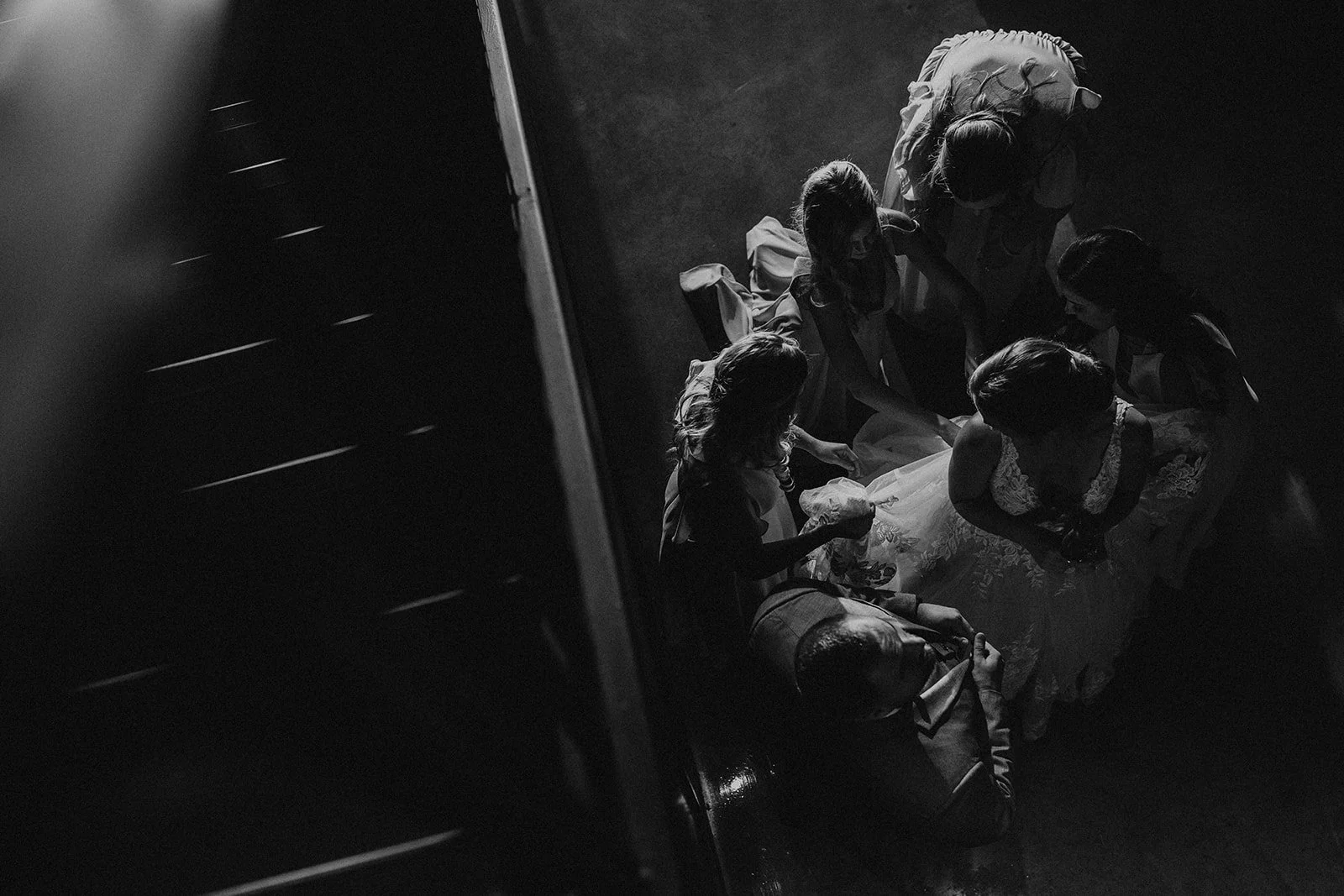 Black and white photo of a group of women, some in wedding dresses, sitting or kneeling on the floor in a circle, viewed from above with a dark background.