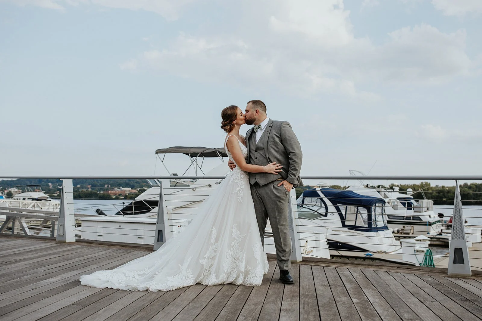 A bride and groom kiss on a dock with boats in the background, the bride in a white lace wedding gown and the groom in a gray suit.