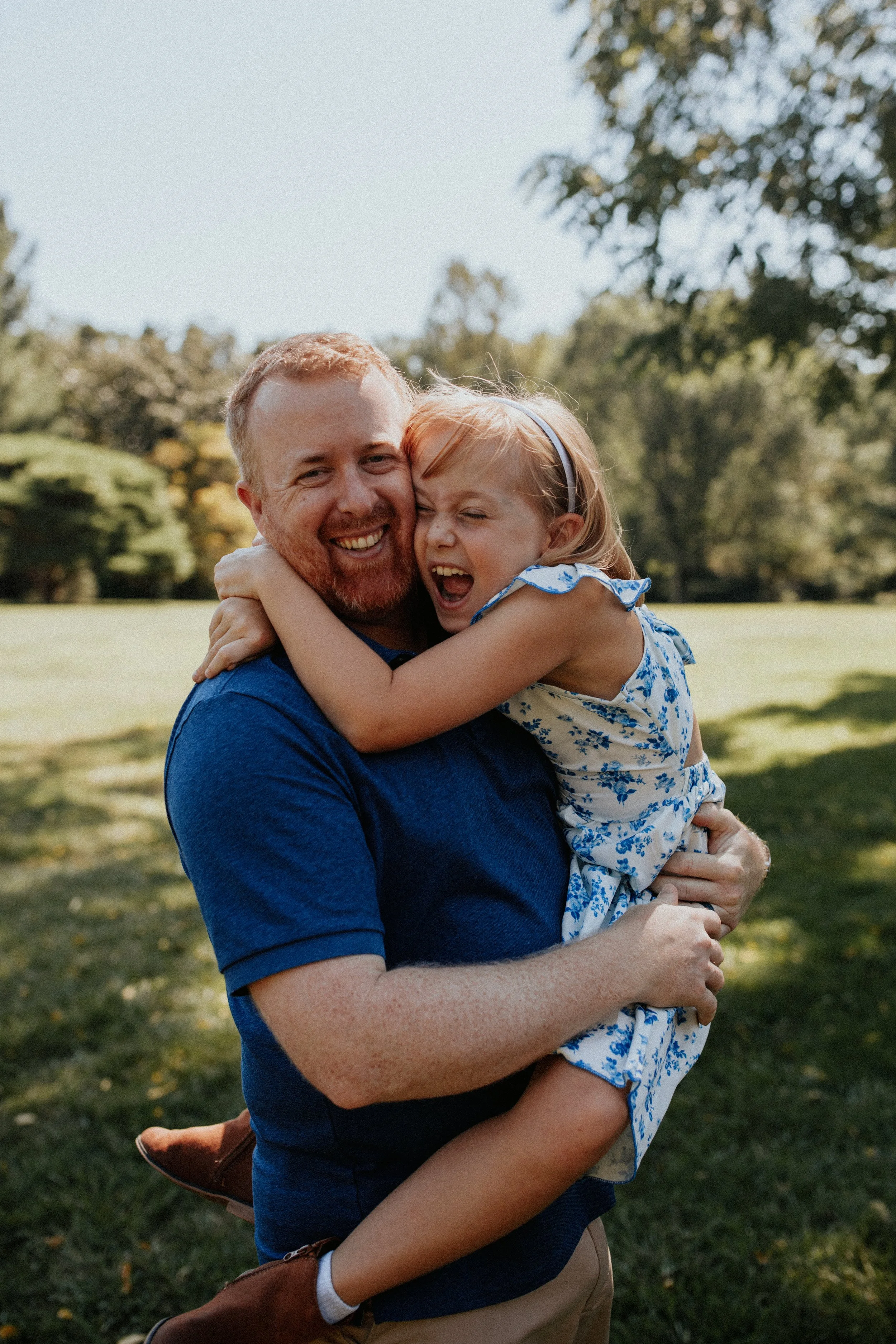 A man with red hair and a beard holding a young girl with red hair and a headband outdoors in a park, both smiling and hugging.