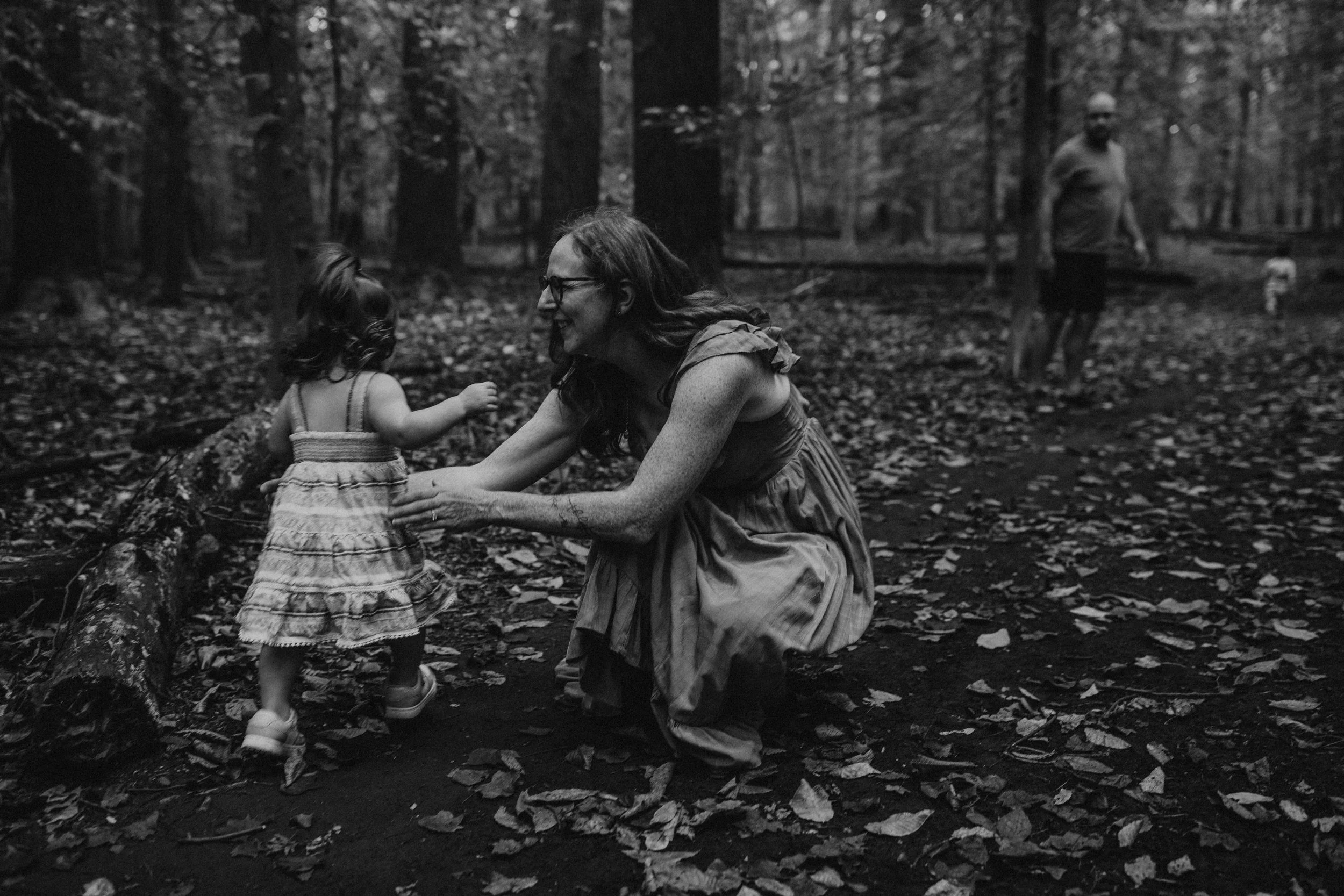 A black and white photo of a woman with glasses, smiling and kneeling on the forest ground, holding hands with a small girl. The girl is wearing a dress and sneakers, and appears to be in the woods surrounded by fallen leaves. In the background, a ma