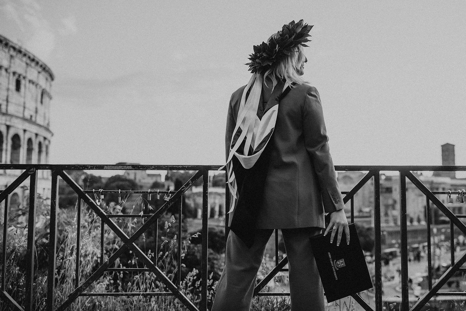 A woman in a blazer and trousers stands on a balcony, wearing a laurel wreath on her head and holding a folder in her right hand, overlooking a cityscape that includes the Colosseum.