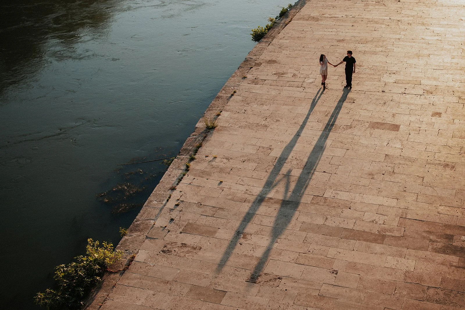 A couple holding hands and walking along a stone-paved riverbank during sunset, with their long shadows cast on the ground.
