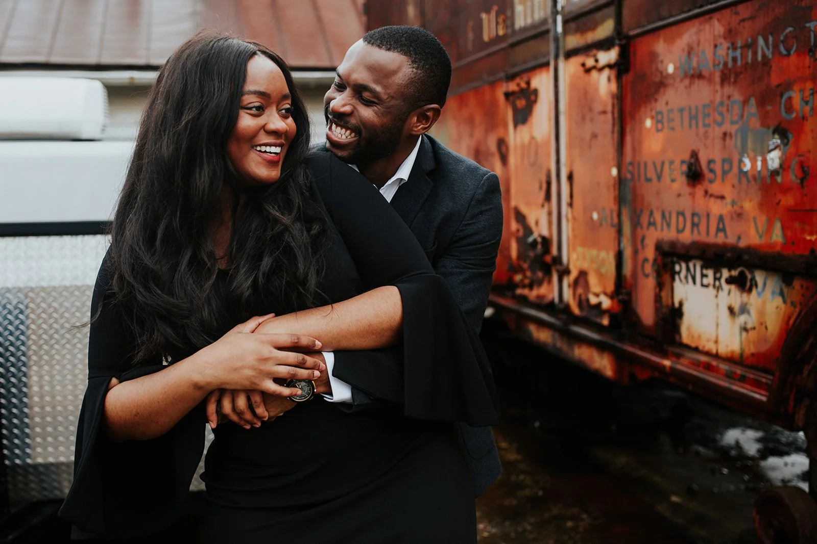 A man and woman smiling and hugging in front of a rusted train car.