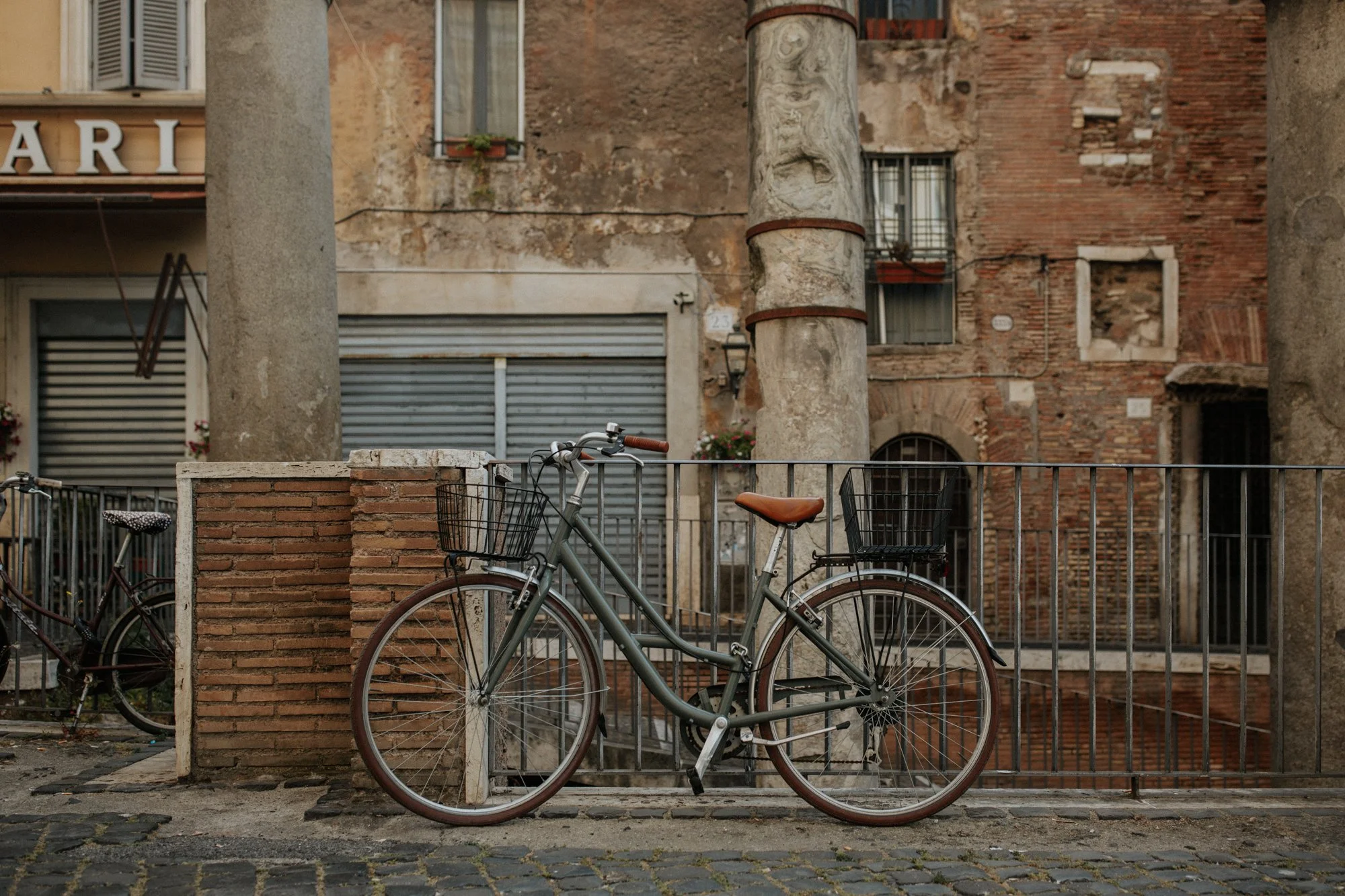 Two bicycles with baskets parked in front of a black metal fence on a cobblestone street, with an old brick and plaster building in the background.