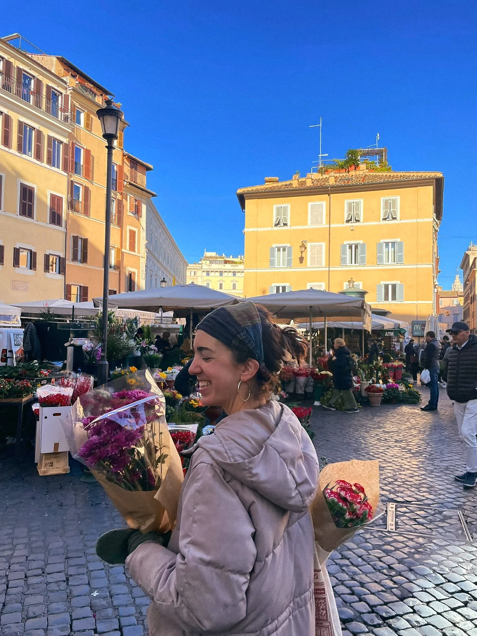 A woman smiling at a flower market on a cobblestone street with colorful buildings and blue sky in the background.