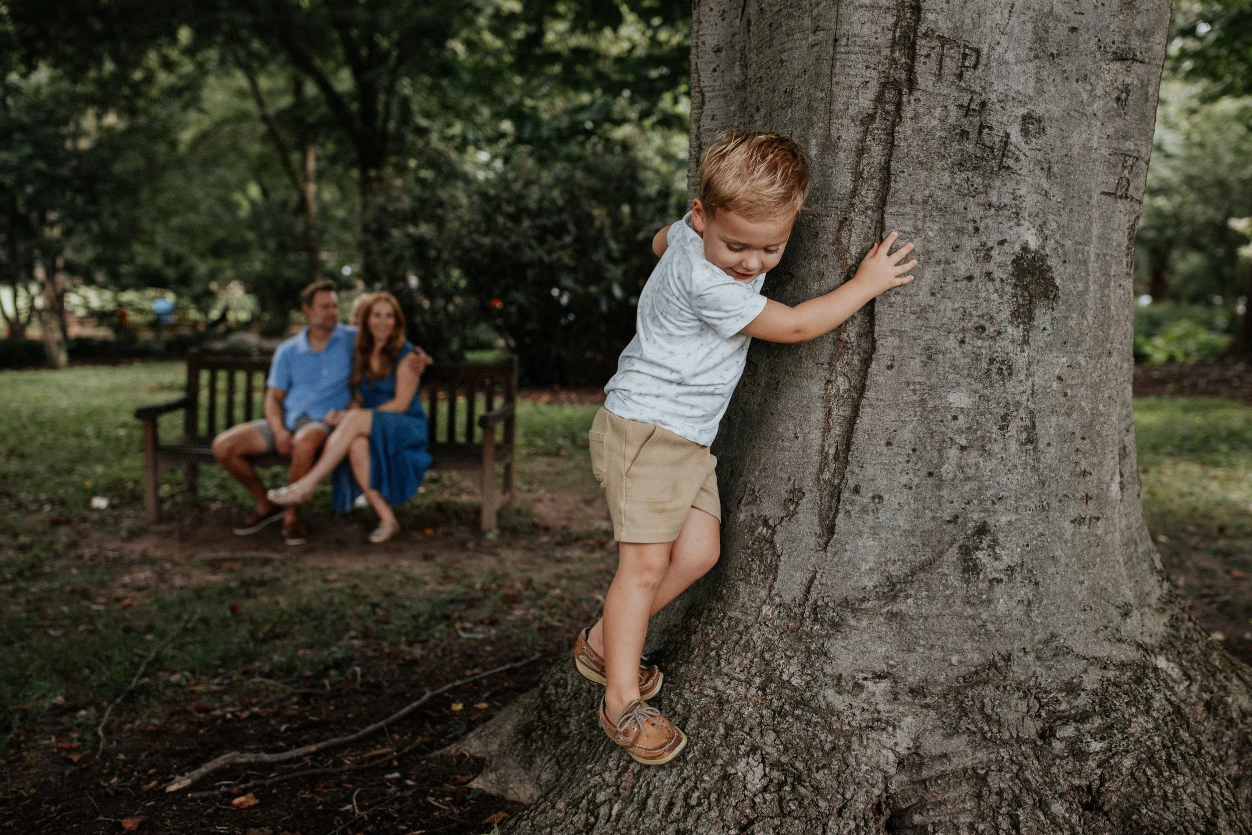 A young boy with blond hair hugging a large tree in a park, with a man and woman sitting on a bench in the background.
