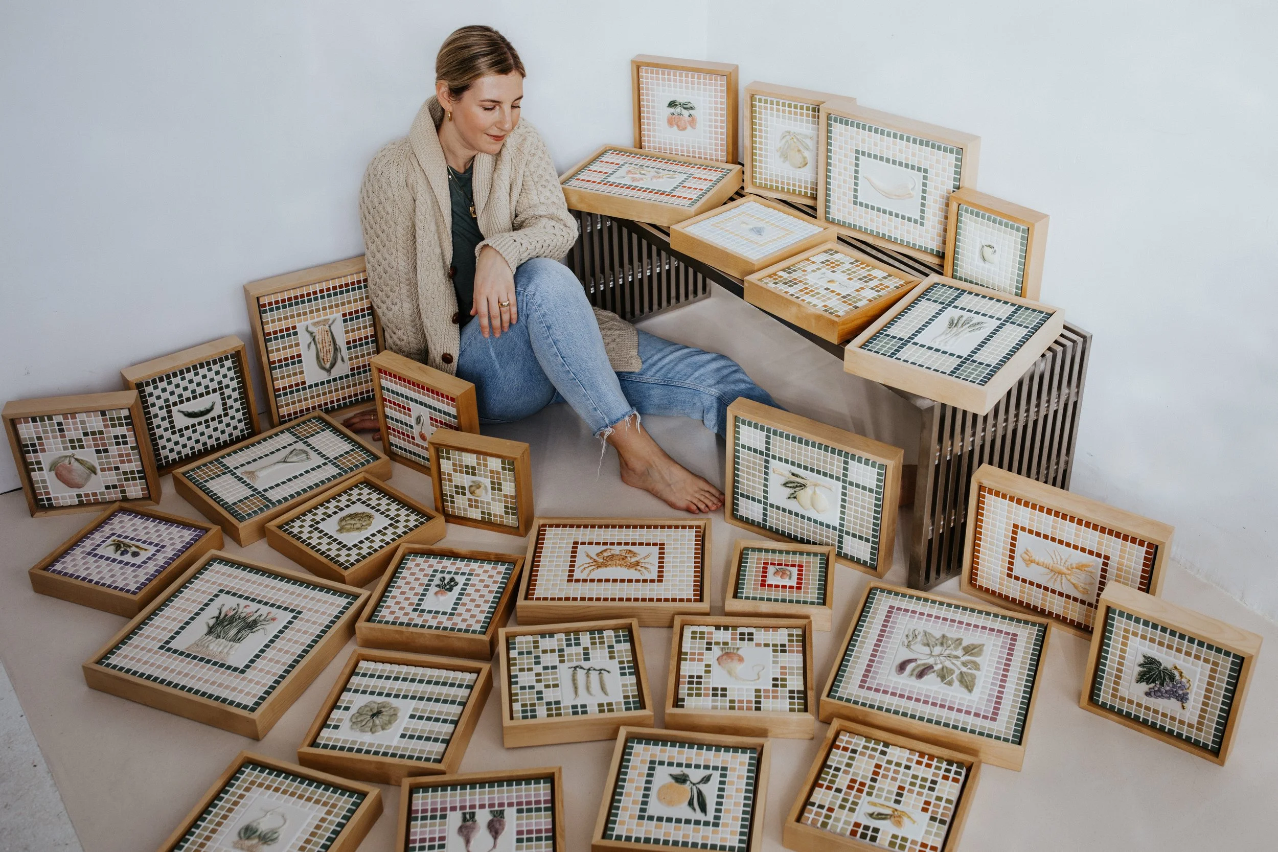A woman sitting on a beige cushion surrounded by framed artwork with various botanical and marine illustrations, including plants, shells, and sea creatures, arranged on the floor and leaning against the wall.