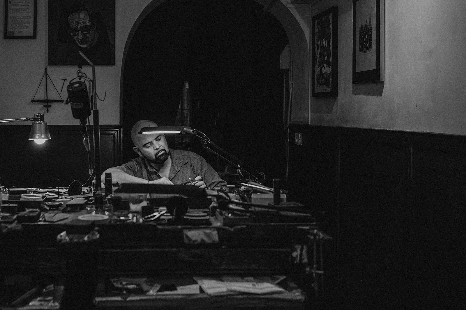 A man with a goatee and bald head sits at a cluttered desk, working under a desk lamp, in a room with dark walls, framed pictures, and artwork.