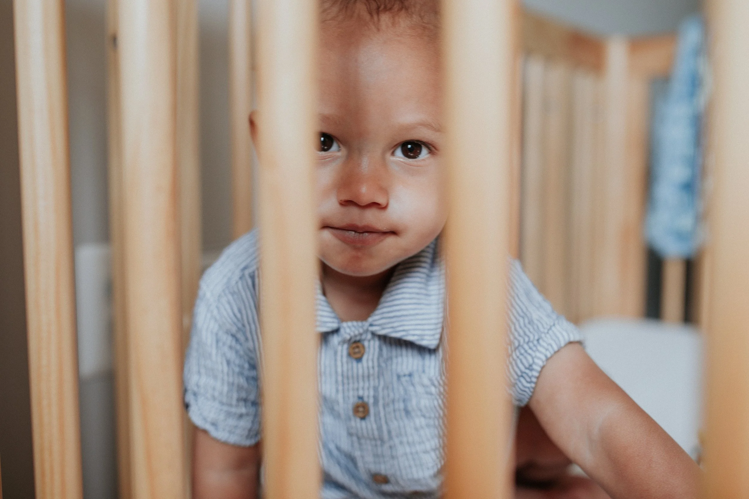 A young child with brown hair and brown eyes looking through wooden bars, wearing a light blue and white striped shirt.