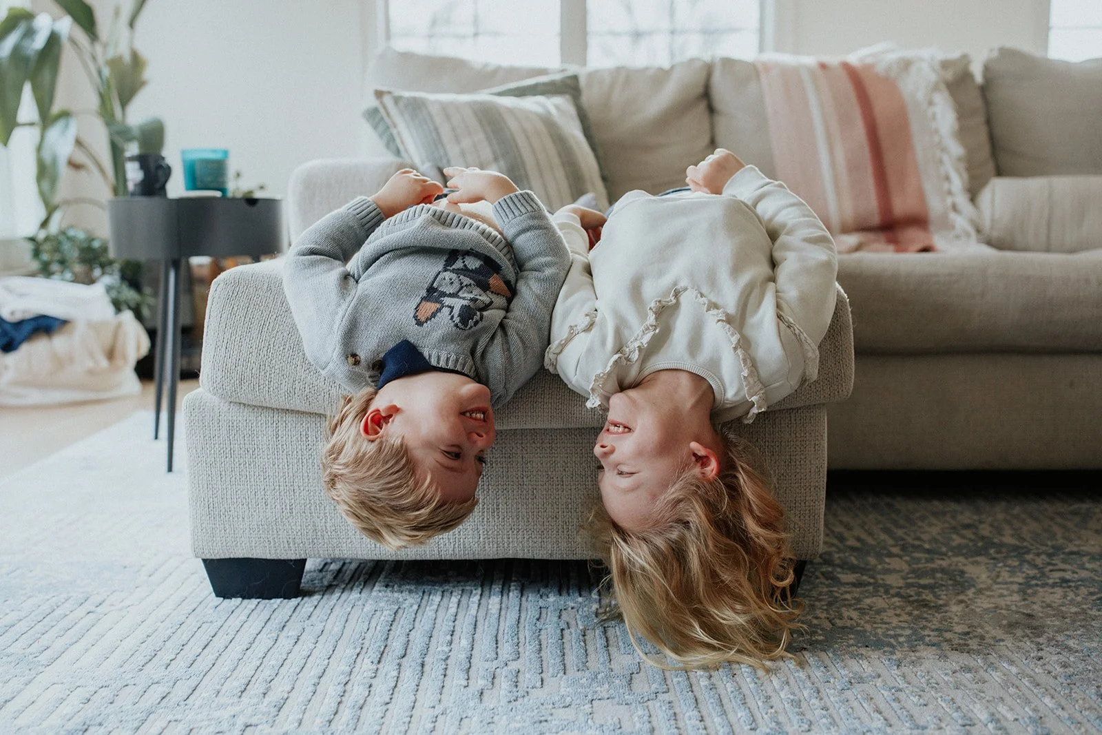 Two children, a boy and a girl, lying upside down on a sofa, smiling and looking at each other in a cozy living room.