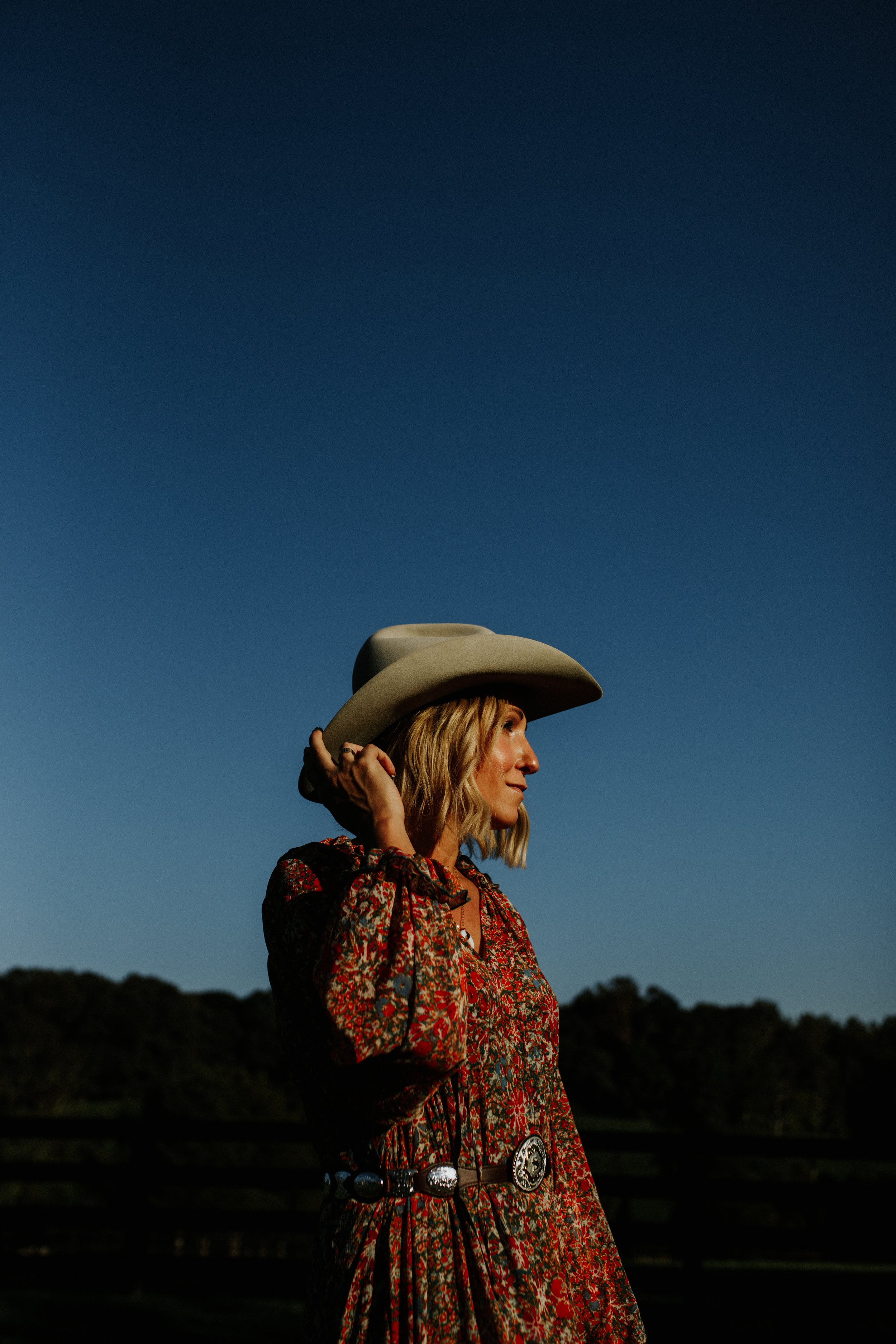 A woman with blonde hair wearing a large cowboy hat and a detailed red patterned dress with a silver belt, standing outdoors during dusk with a dark blue sky and trees in the background.