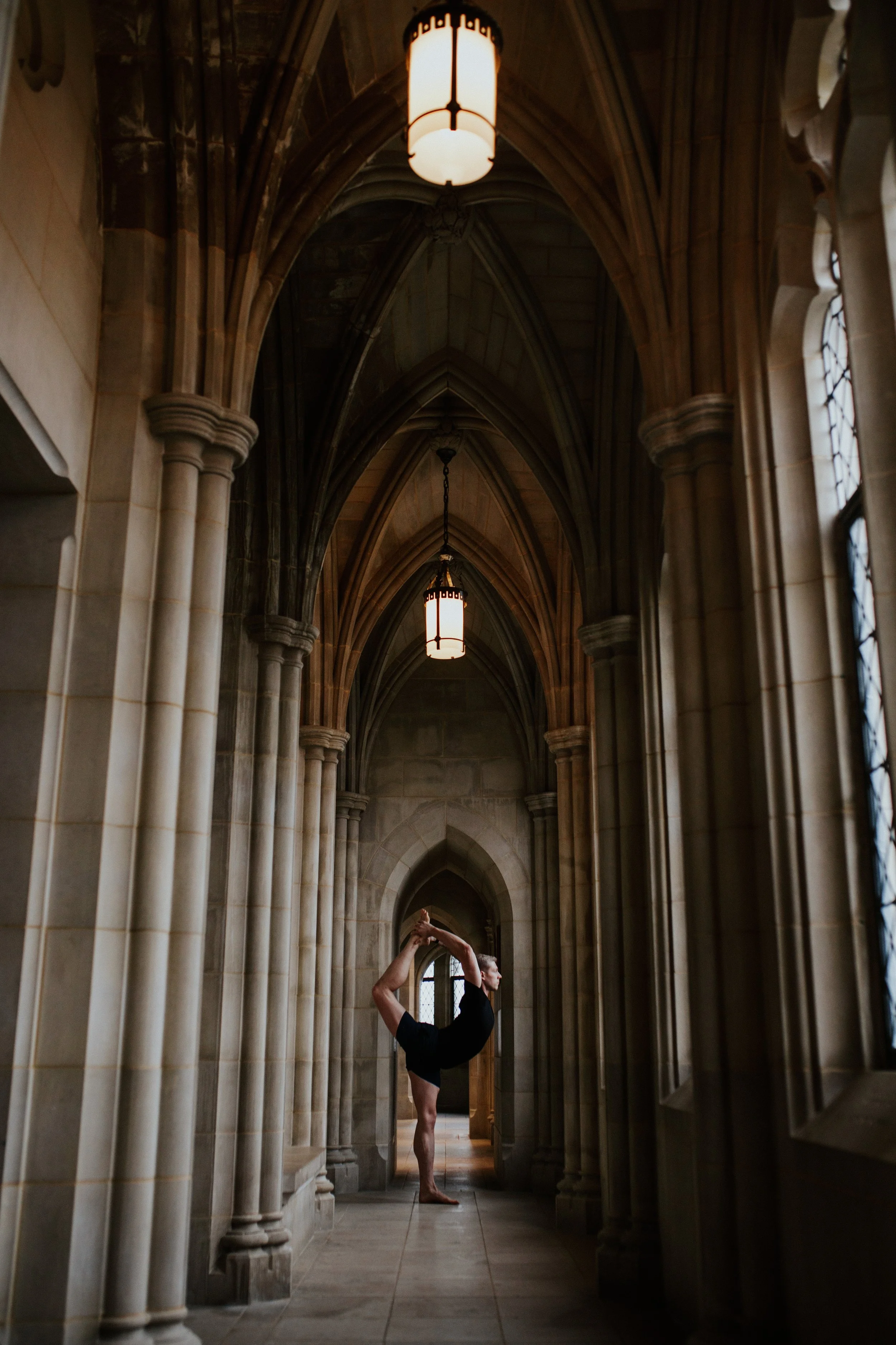 A man practicing the yoga pose known as dancer's pose in a corridor of a historic stone Gothic church, featuring arches and hanging lanterns.