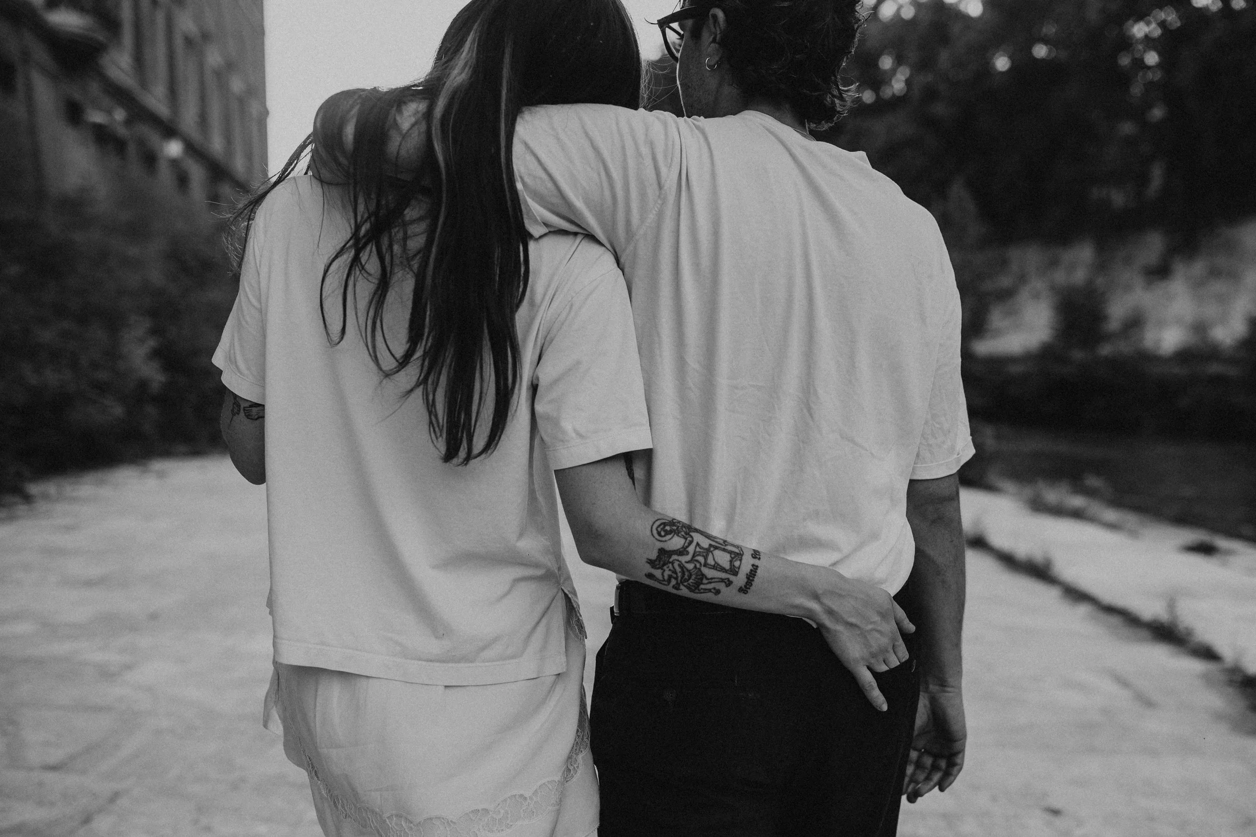 A black and white photo of two people standing close together outdoors, with one person's arm around the other's shoulder. The person on the left has long dark hair and tattoos on their arm, wearing a short-sleeved shirt. The person on the right has 