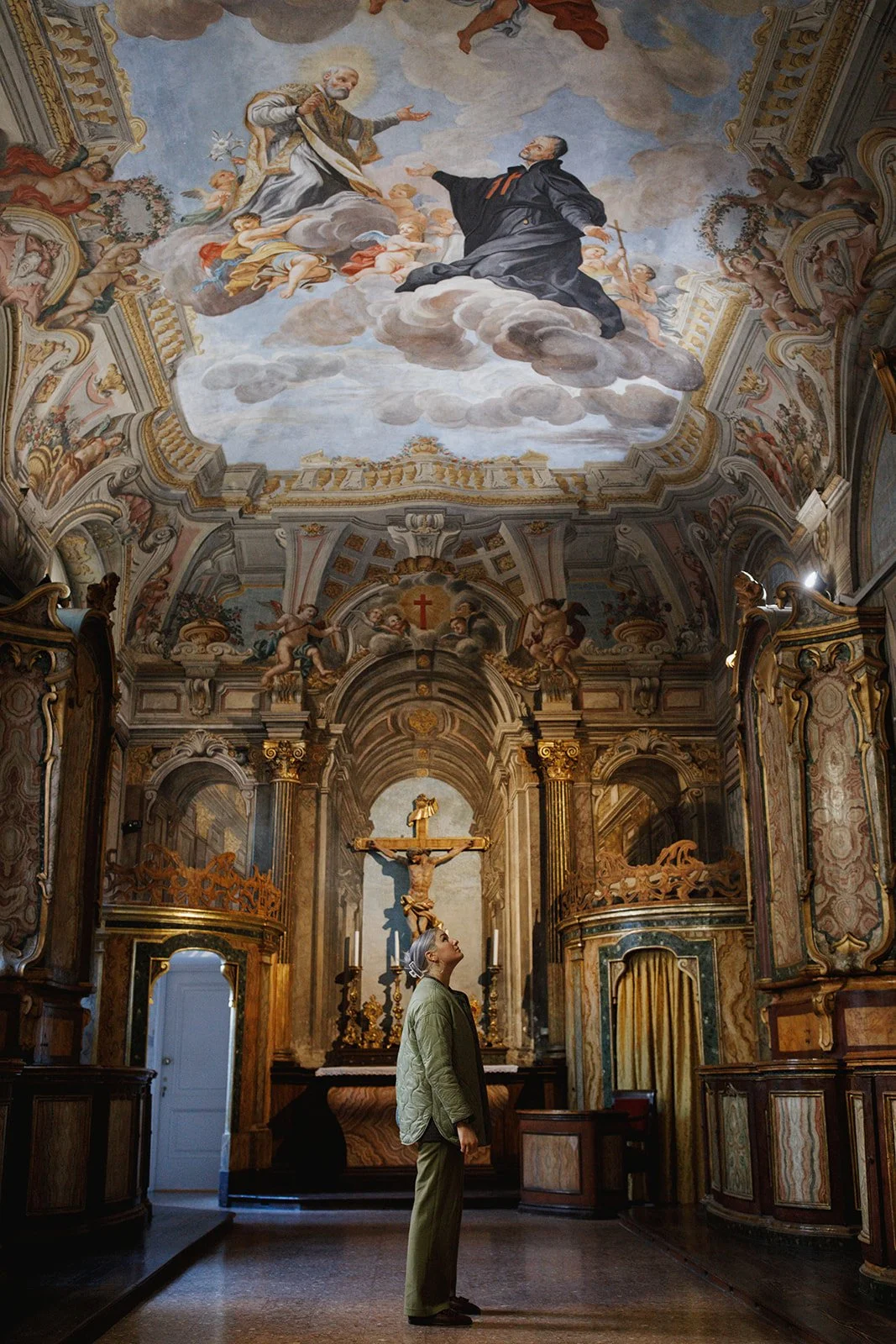 A person standing inside a richly decorated Catholic church, looking up at a large ornate altar with a crucifix, elaborate woodwork, and gold accents. The ceiling features a colorful fresco painting of religious figures in the sky with clouds.