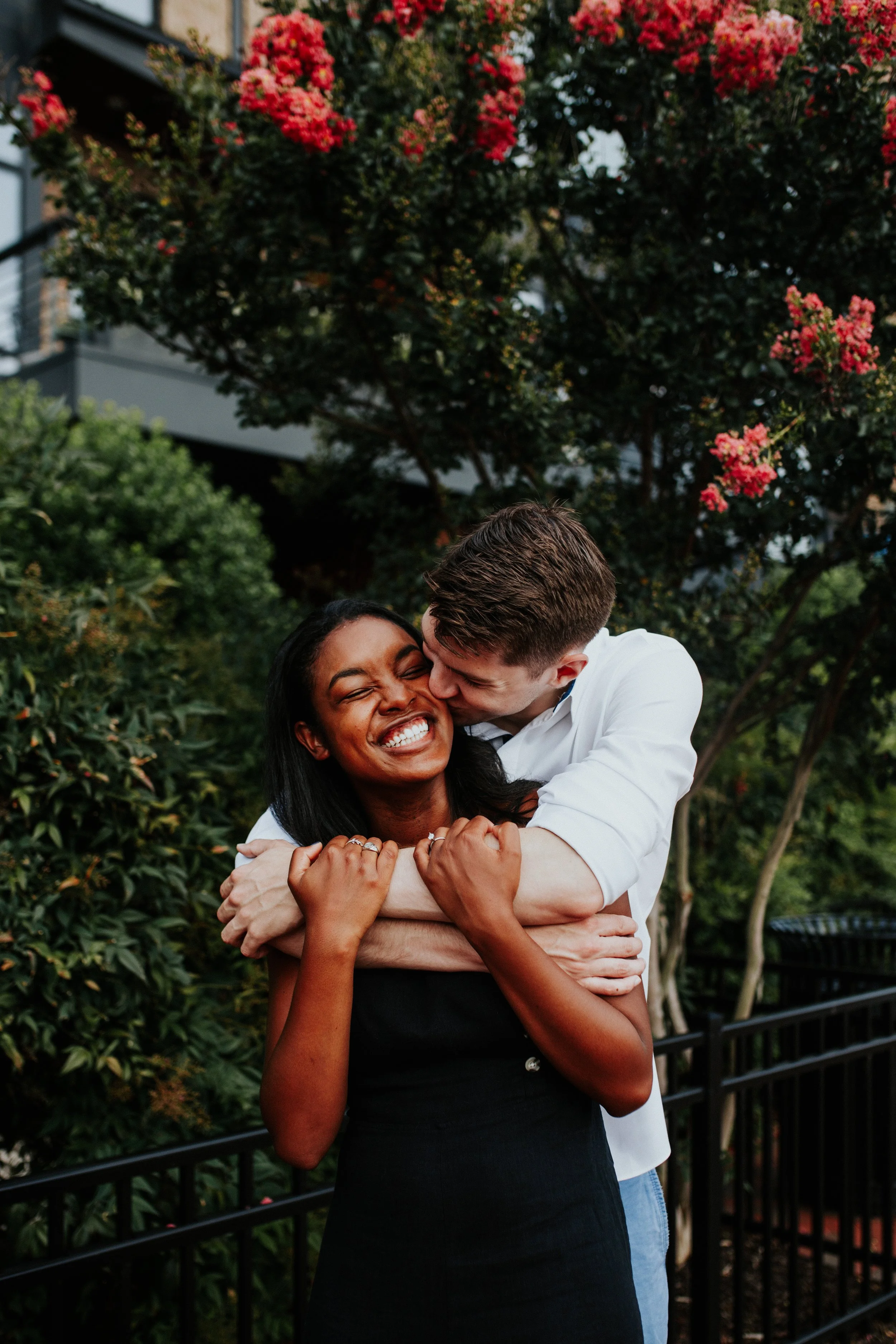 A man and woman embracing and smiling, with the man kissing the woman on the cheek, in an outdoor setting with green bushes and pink flowering trees in the background.