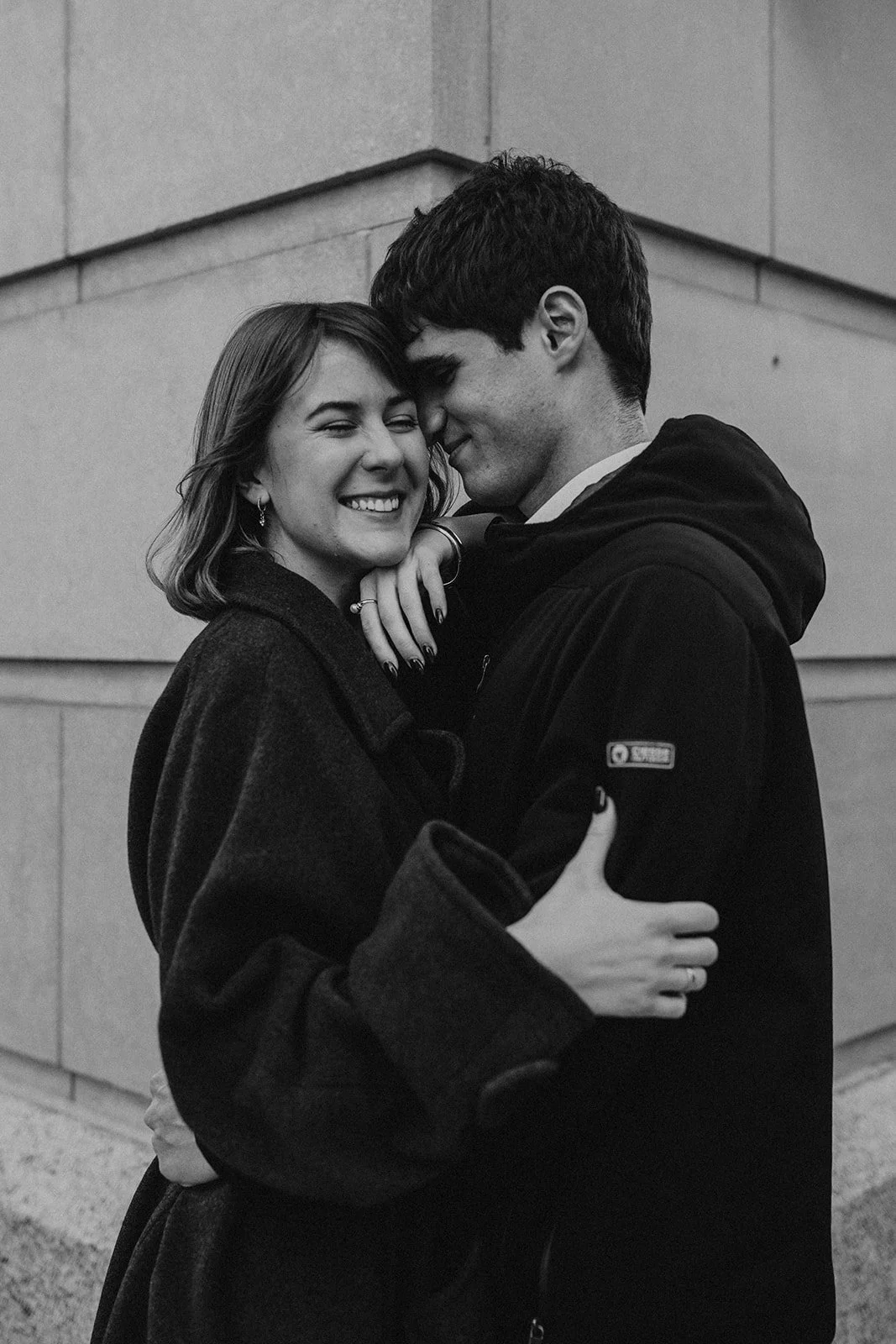 A black-and-white photo of a smiling woman and man embracing outdoors, with stone wall background.