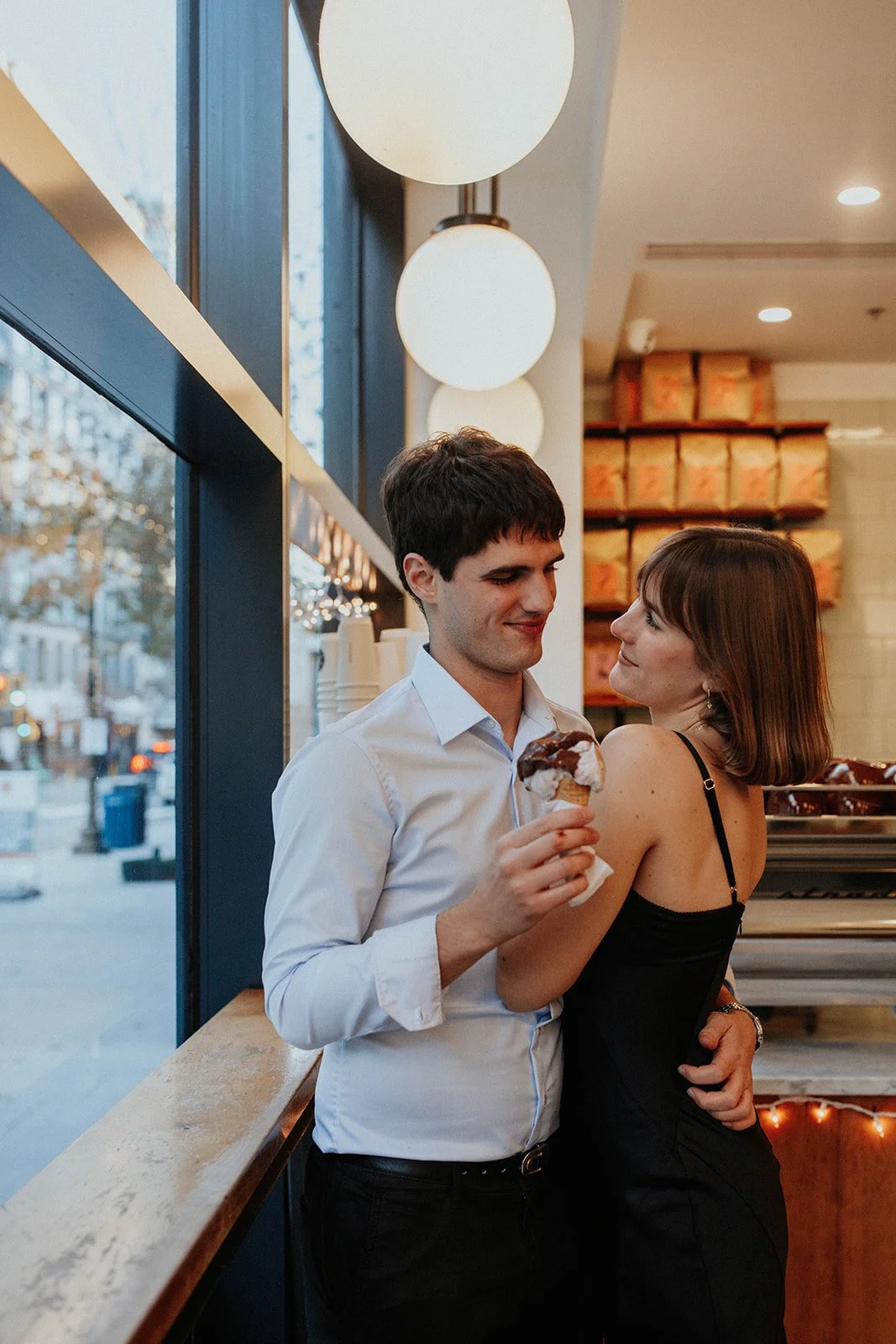 A man and woman in a cafe or ice cream shop, with the man holding an ice cream cone with chocolate topping, both smiling and looking at each other, near a window with street outside view.
