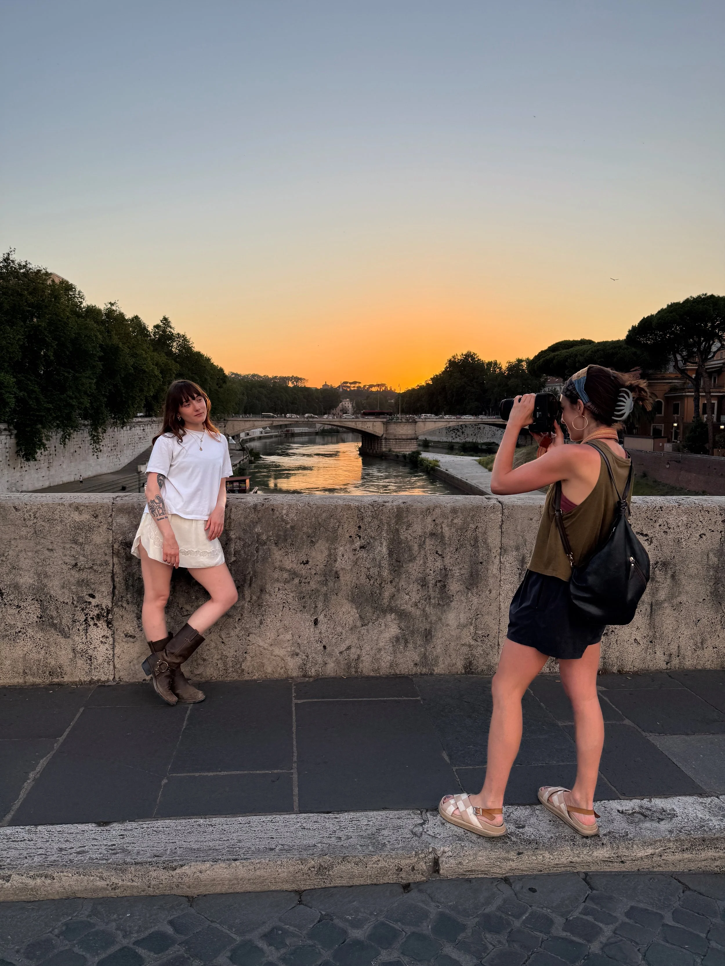 Two women on a bridge during sunset, one in a white t-shirt and cream skirt with tattoos, the other taking a photograph, wearing a green top, shorts, and sandals, with trees and buildings in the background.